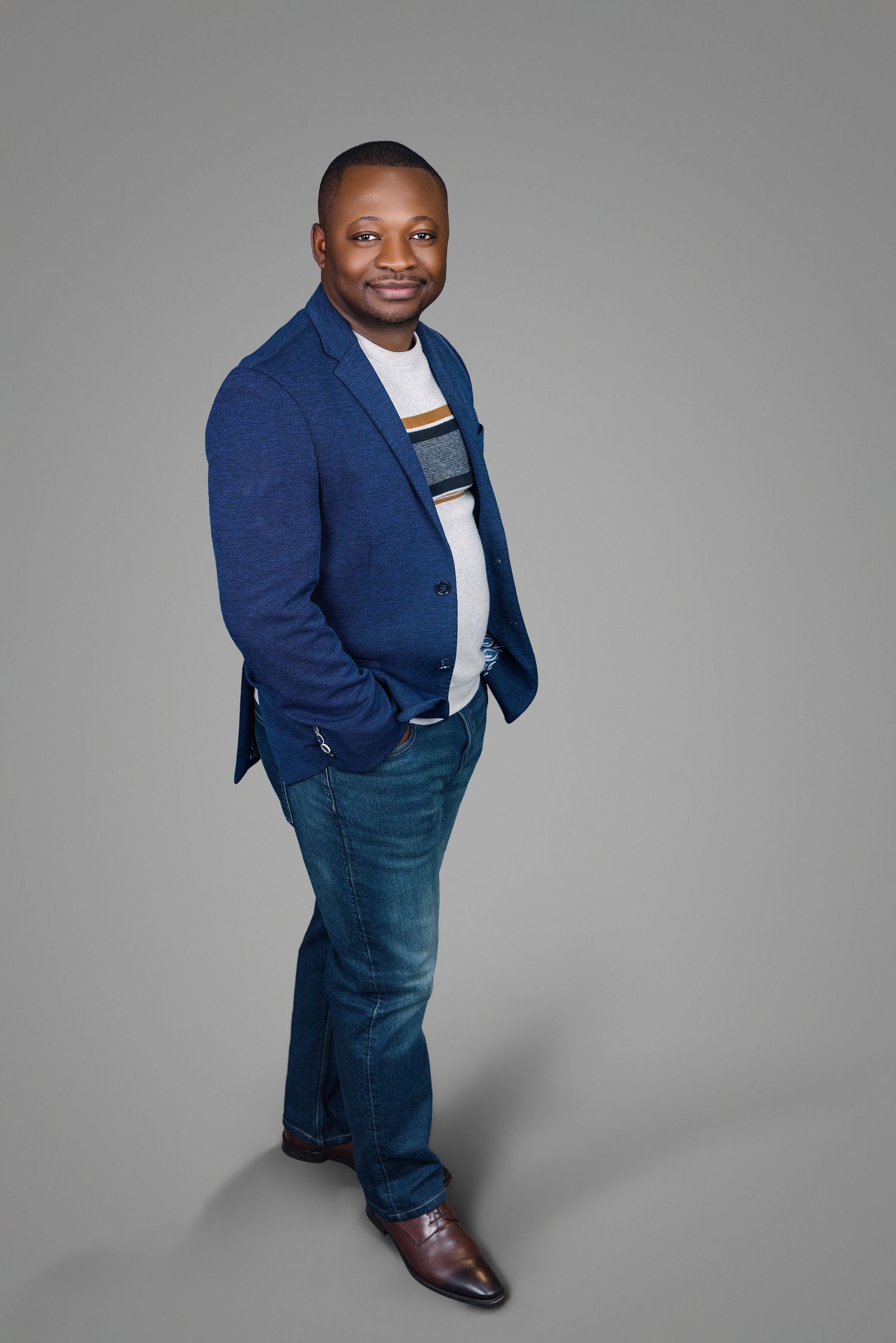 Cheerful Black man wearing blue blazer and striped shirt stands with hands in pockets on gray backdrop.