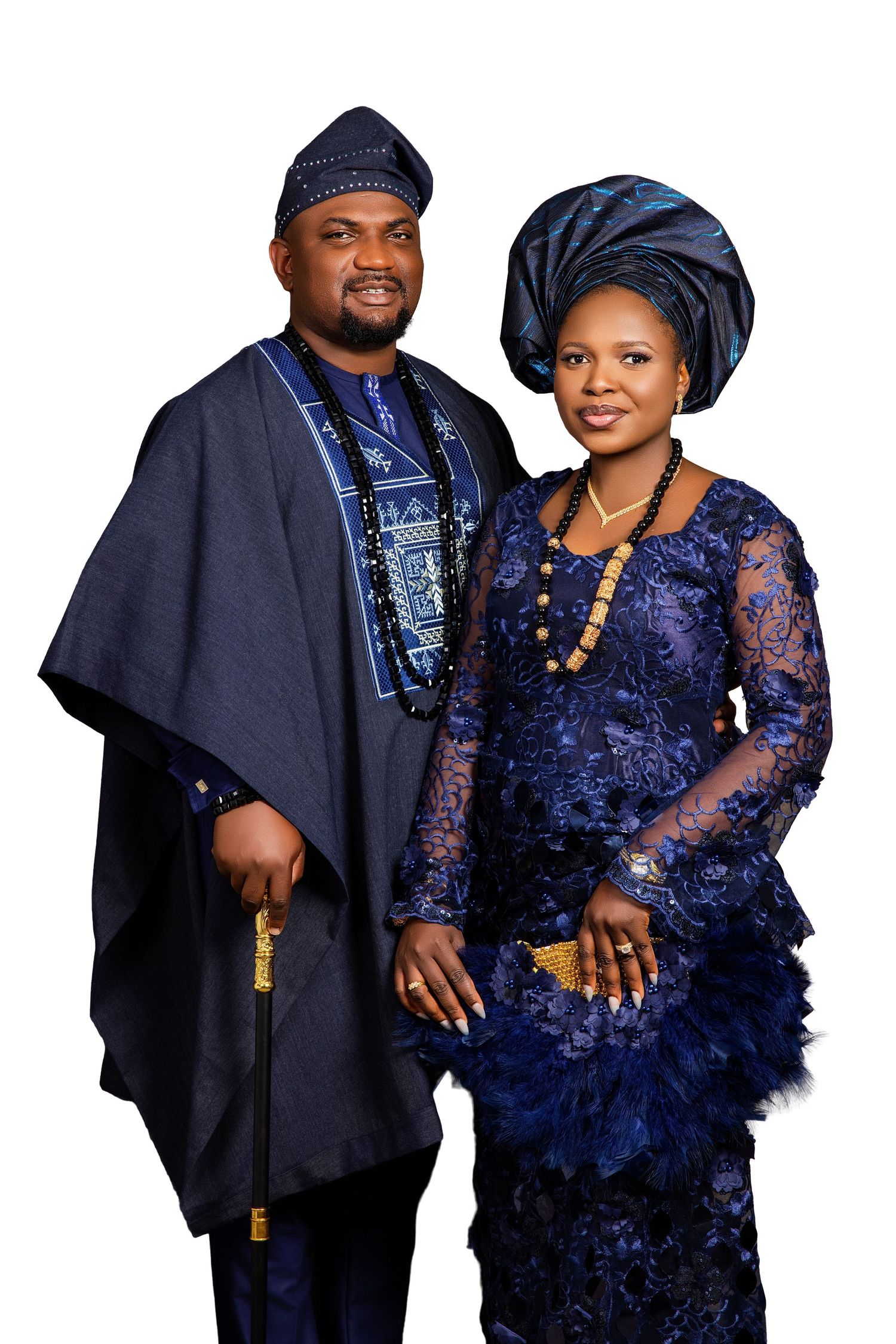 Nigerian couple in matching navy blue traditional Yoruba attire, agbada and lace gele, posing formally against white background.
