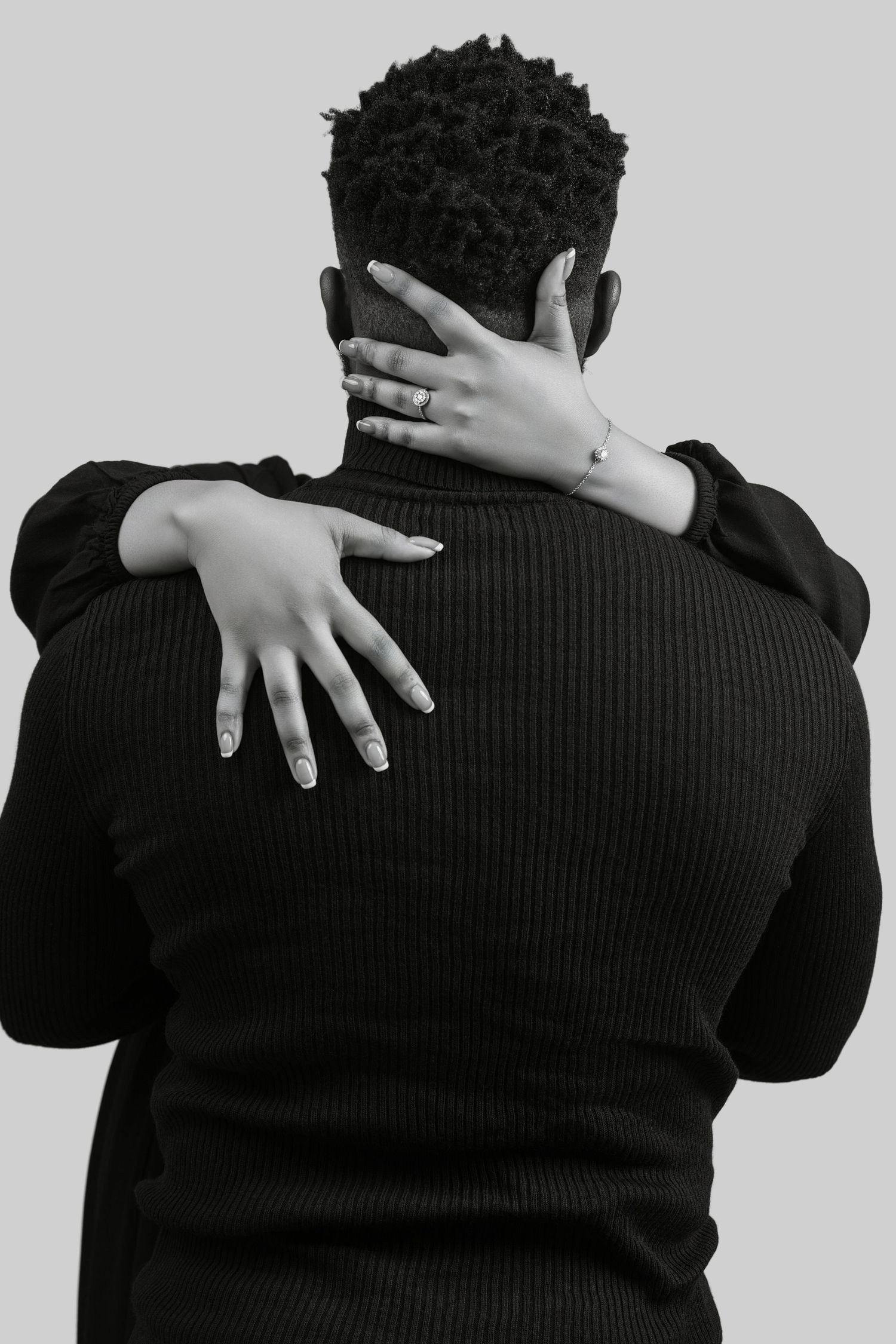 Black and white photo of a woman embracing a man from behind, her hands adorned with rings and bracelet.
