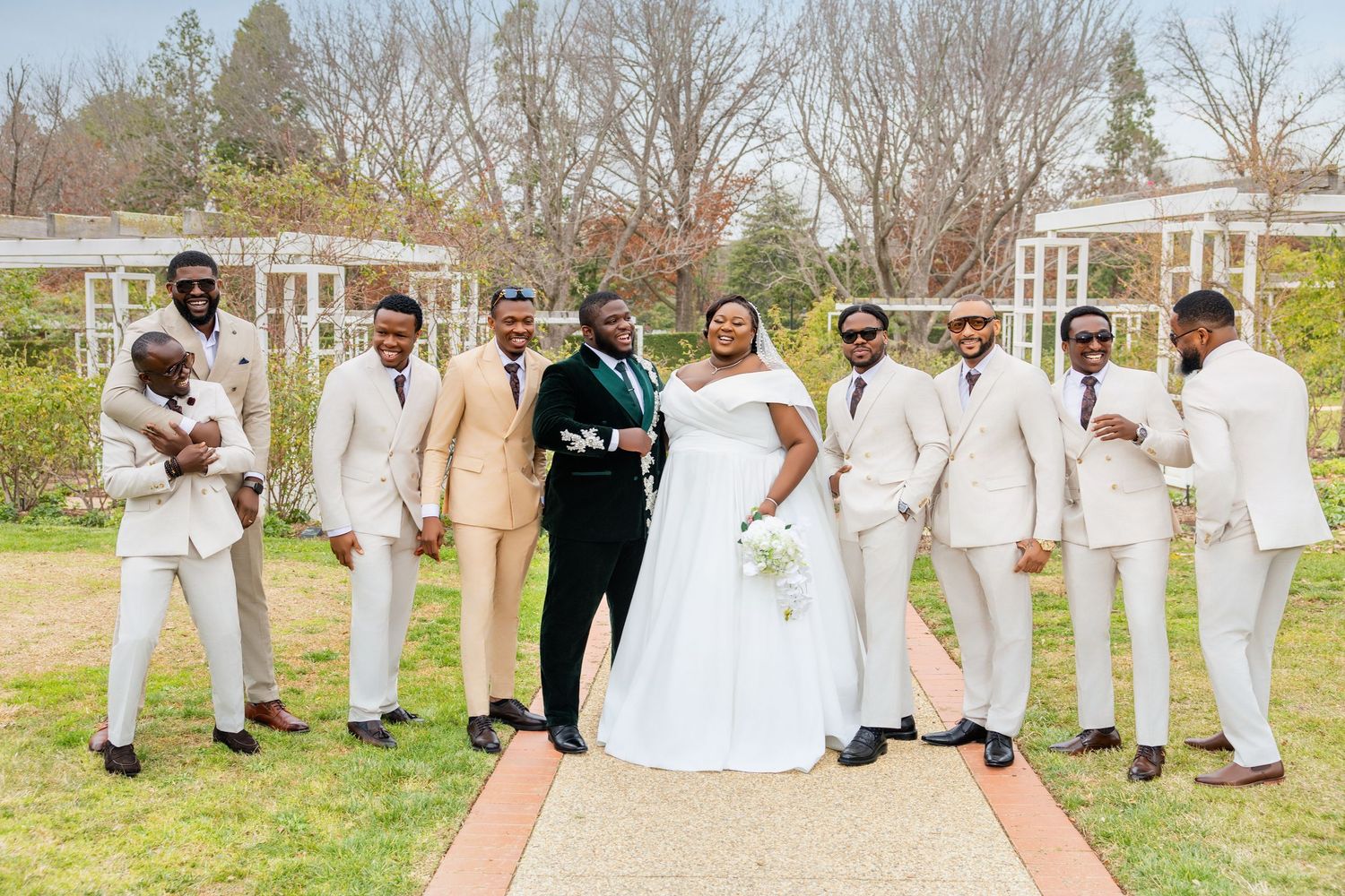 A bride and groom pose outdoors with their wedding party in beige suits and green accents.