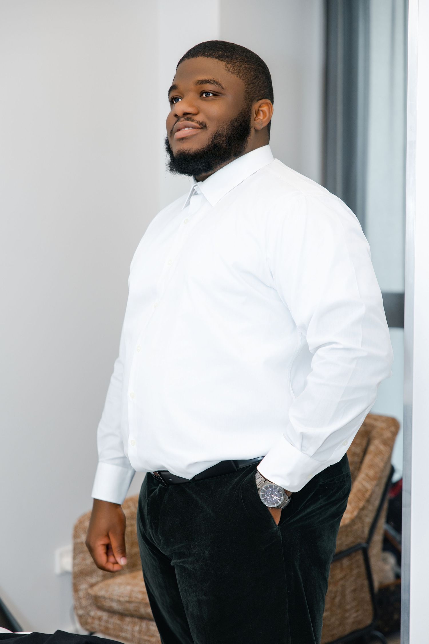 A confident Black man in a white dress shirt and dark pants smiles while standing near a window indoors.