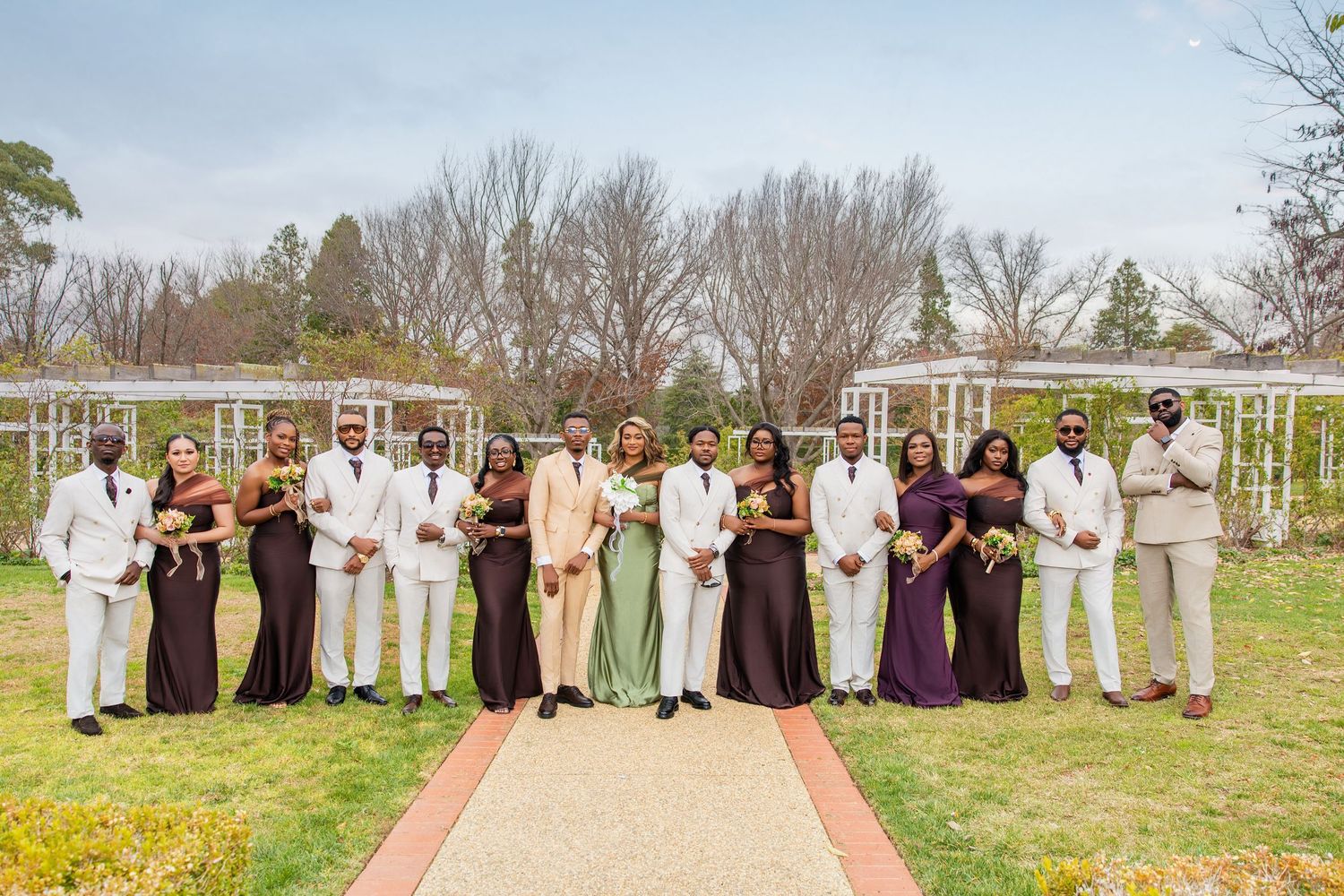Wedding party in purple bridesmaid dresses and white suits posing outdoors in garden with pergola backdrop.