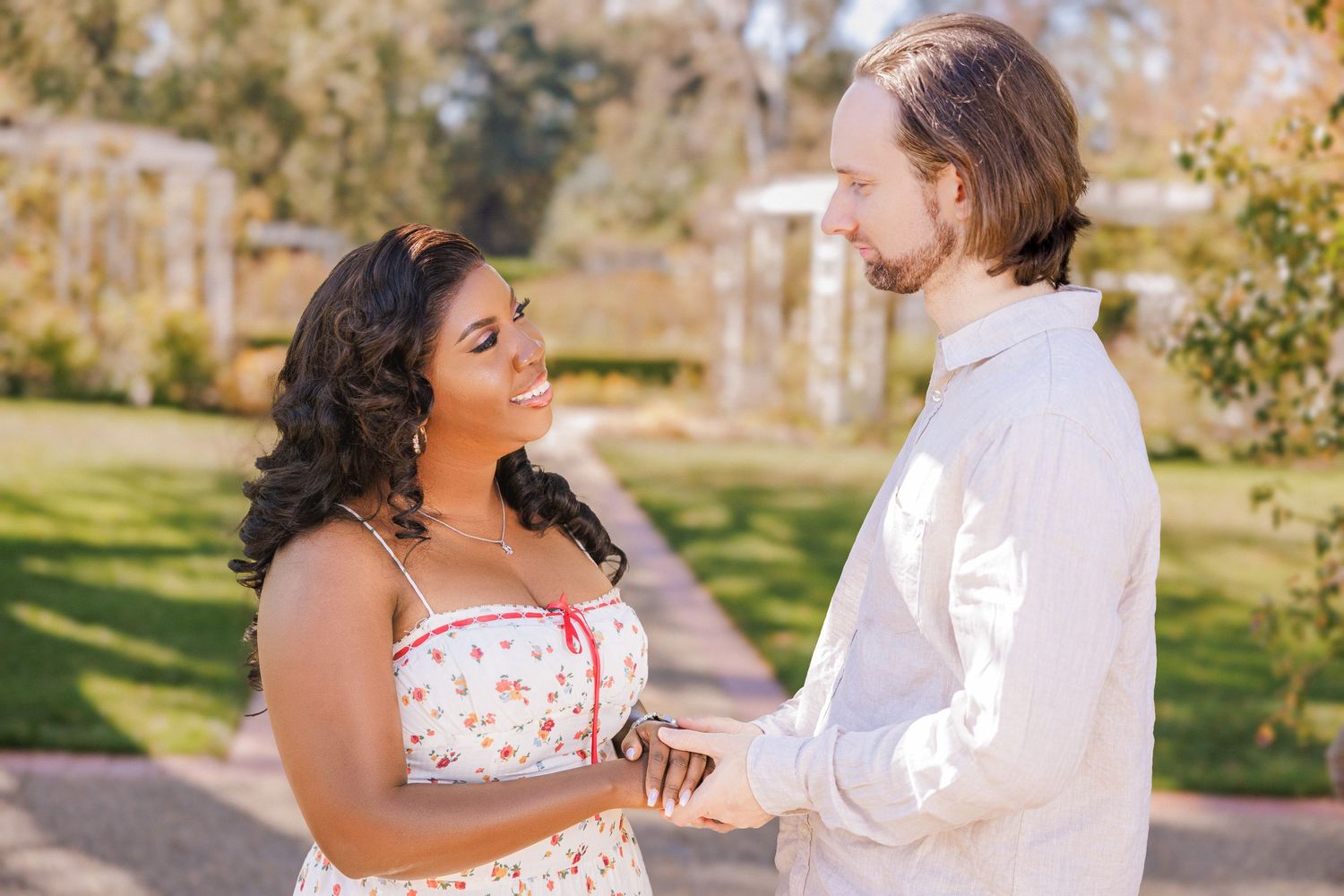 A joyful couple holds hands and smiles at each other in a sunlit garden, dressed in casual summer attire.