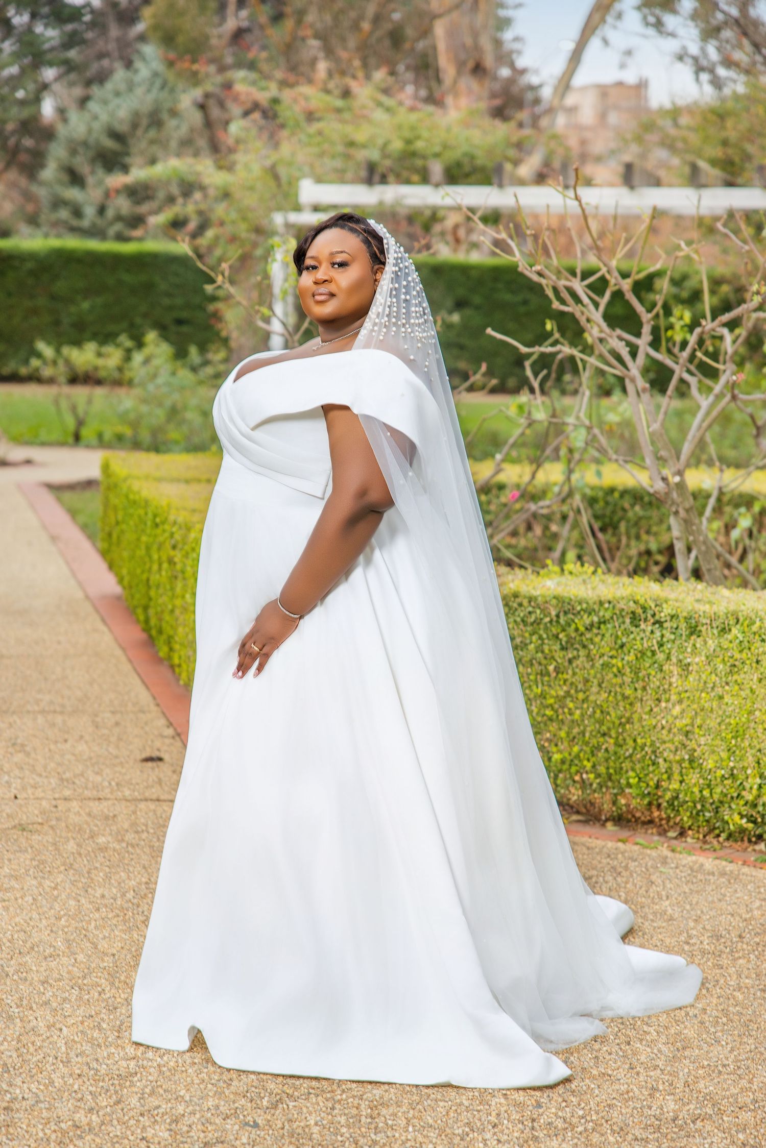 Beautiful bride in flowing white off-shoulder wedding gown with embellished veil posing in lush garden setting.