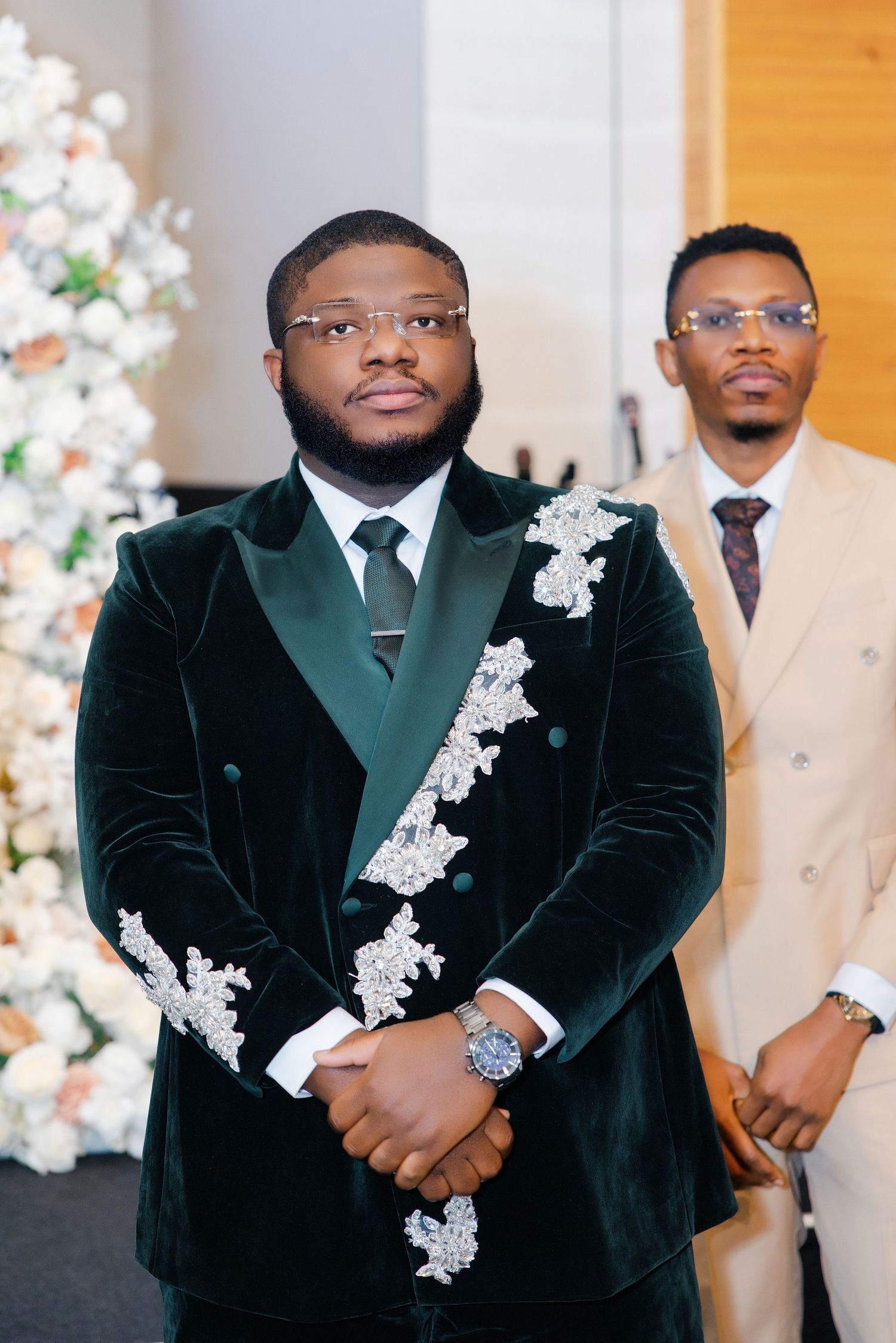 Two men in elegant formal attire pose at a wedding, one in a green velvet embroidered tuxedo.