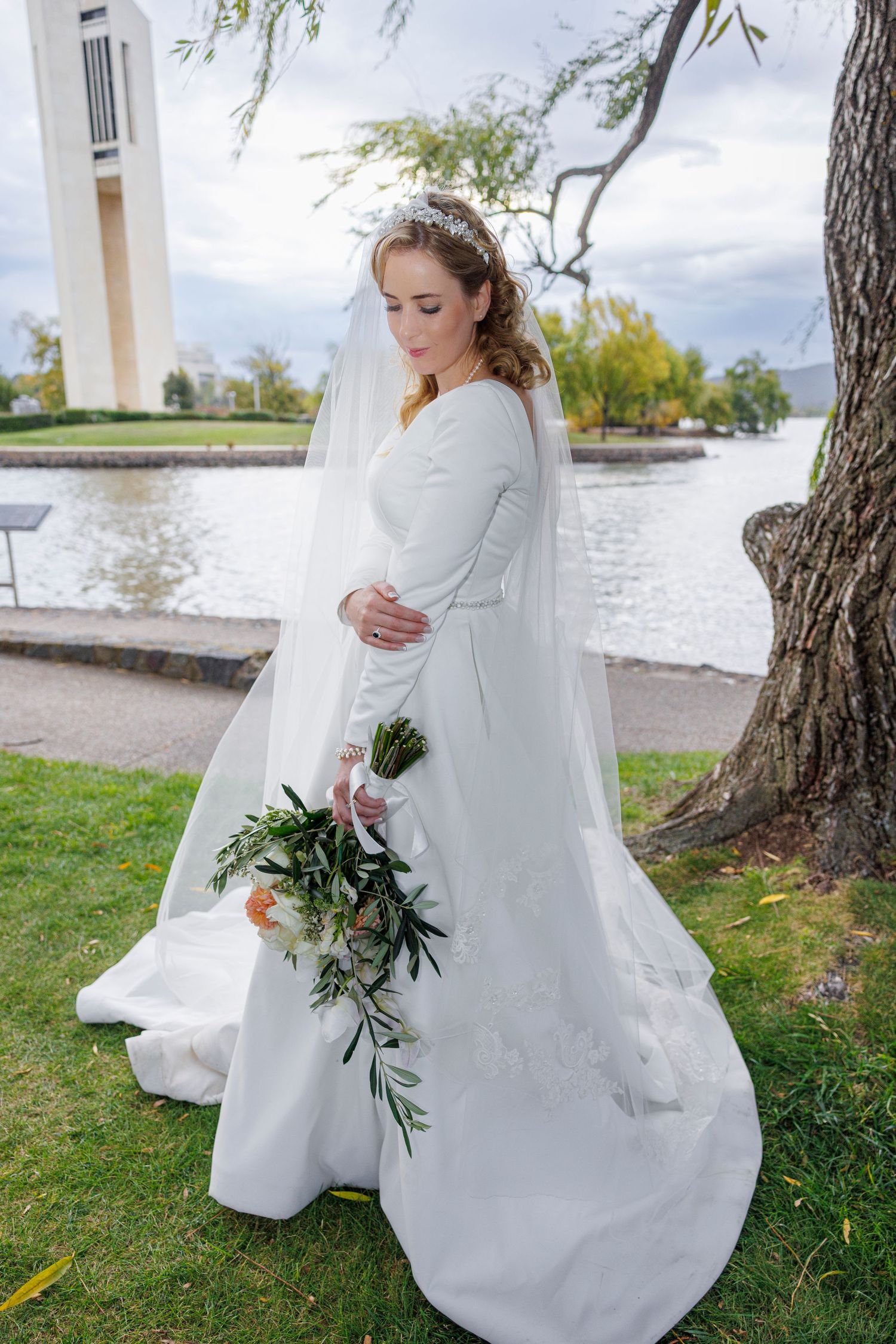Bride in white gown with long veil holding bouquet near a lakeside monument.