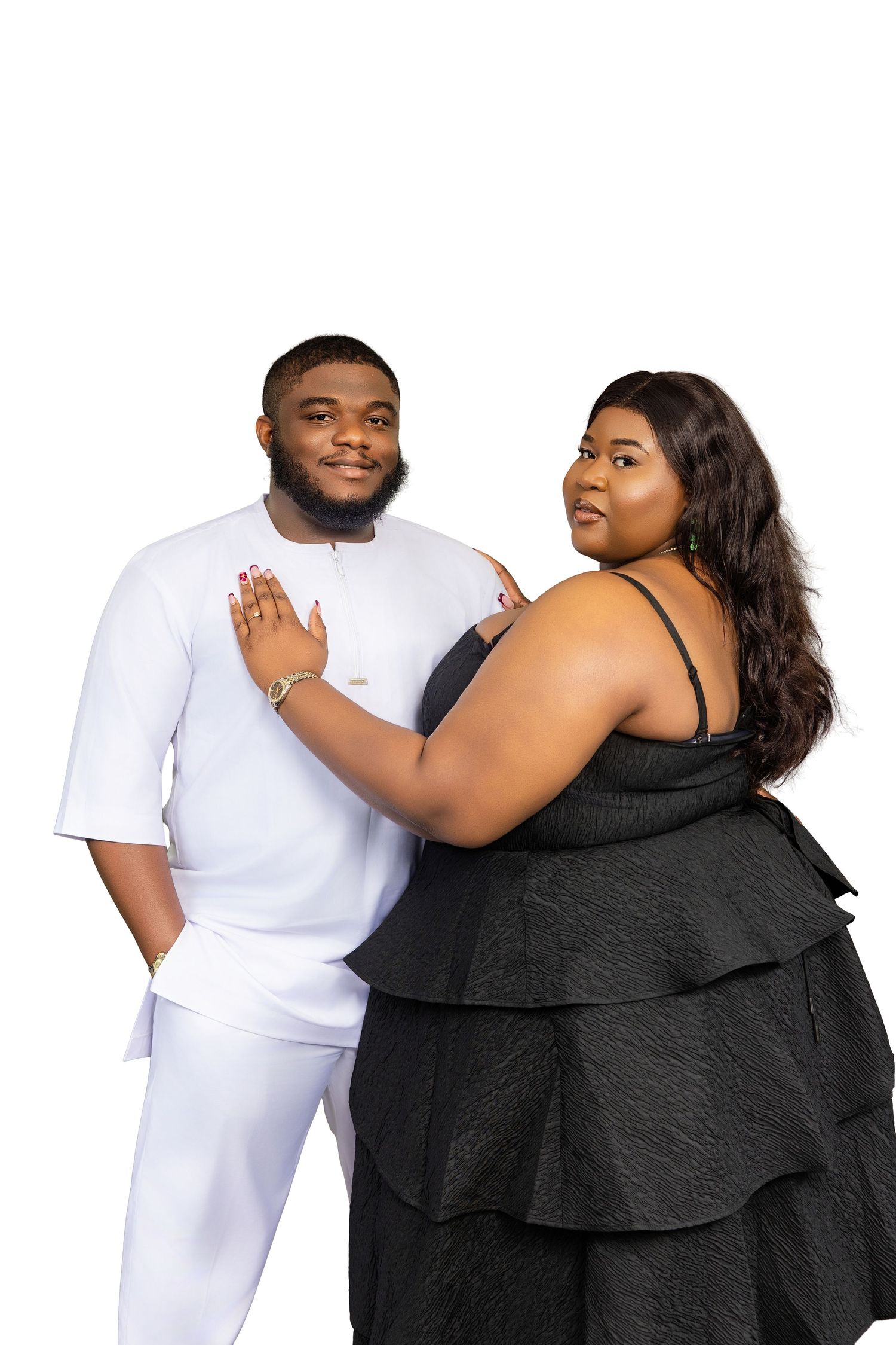 A couple in formal attire poses together, man in white outfit and woman in black dress, on white backdrop.