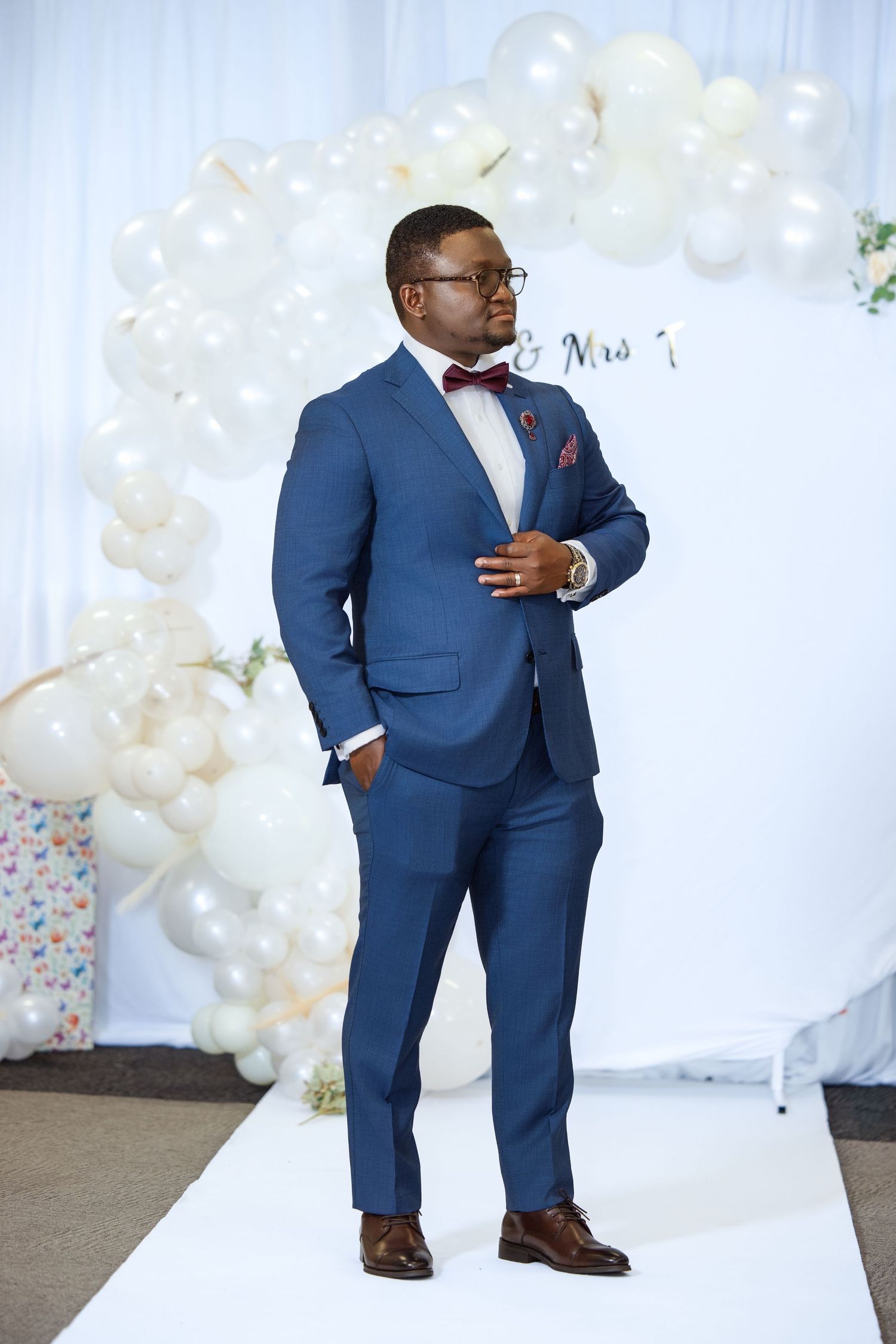 Elegant man in blue suit with bow tie poses at wedding reception with balloon arch backdrop.