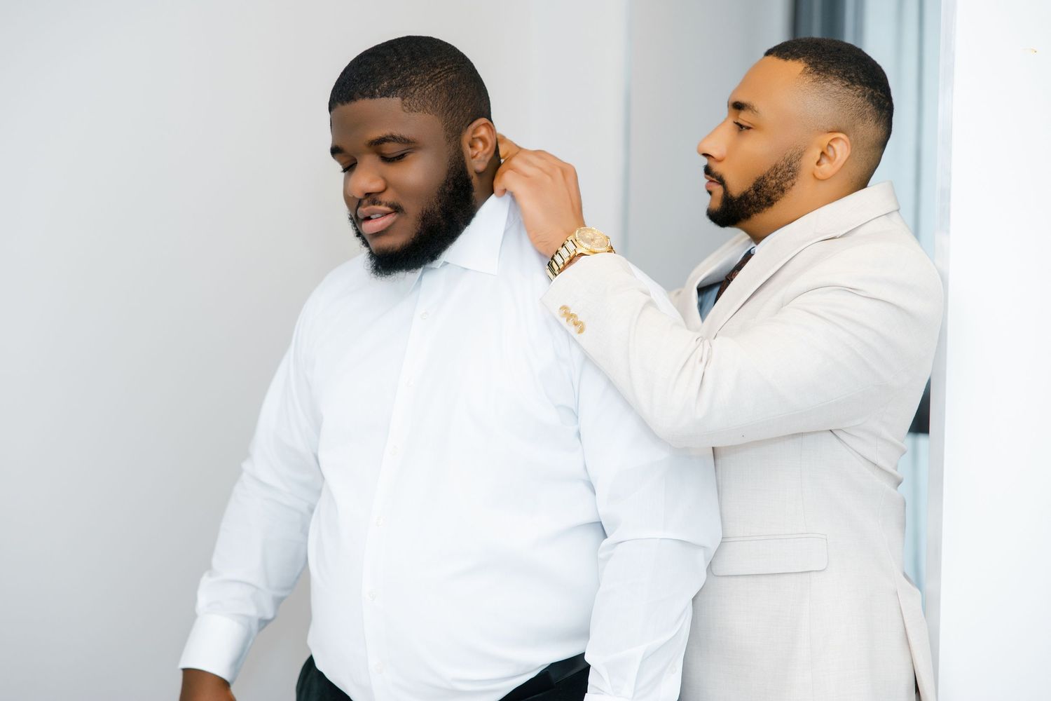 A groomsman in a white suit adjusts the collar of a man in a white dress shirt before a wedding.