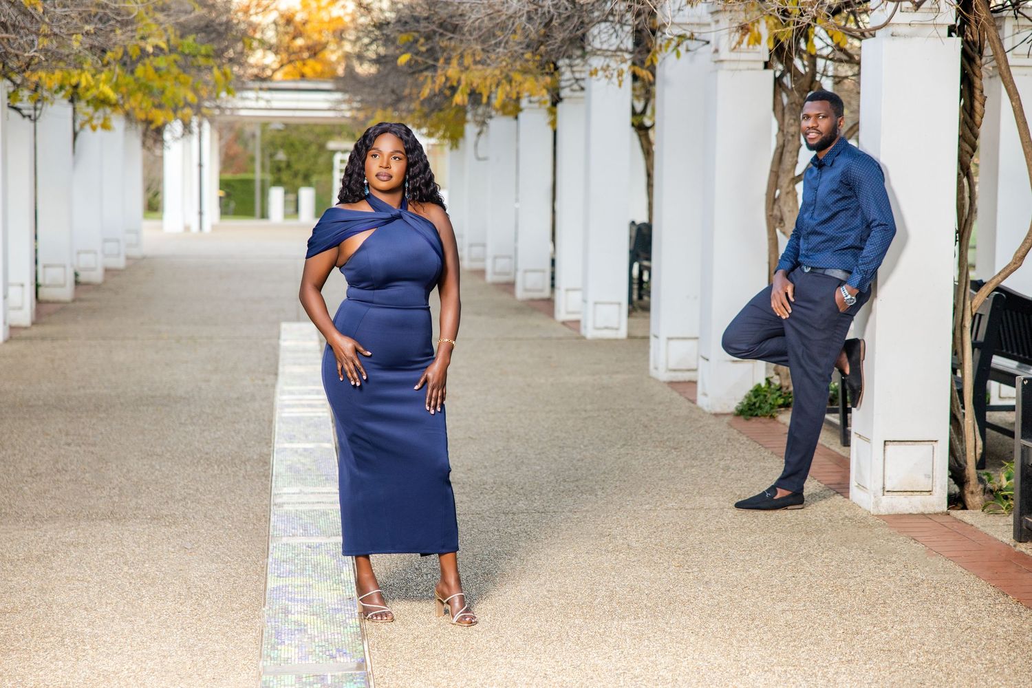 Elegant couple in matching navy blue outfits posing in a white columned outdoor walkway with autumn trees.