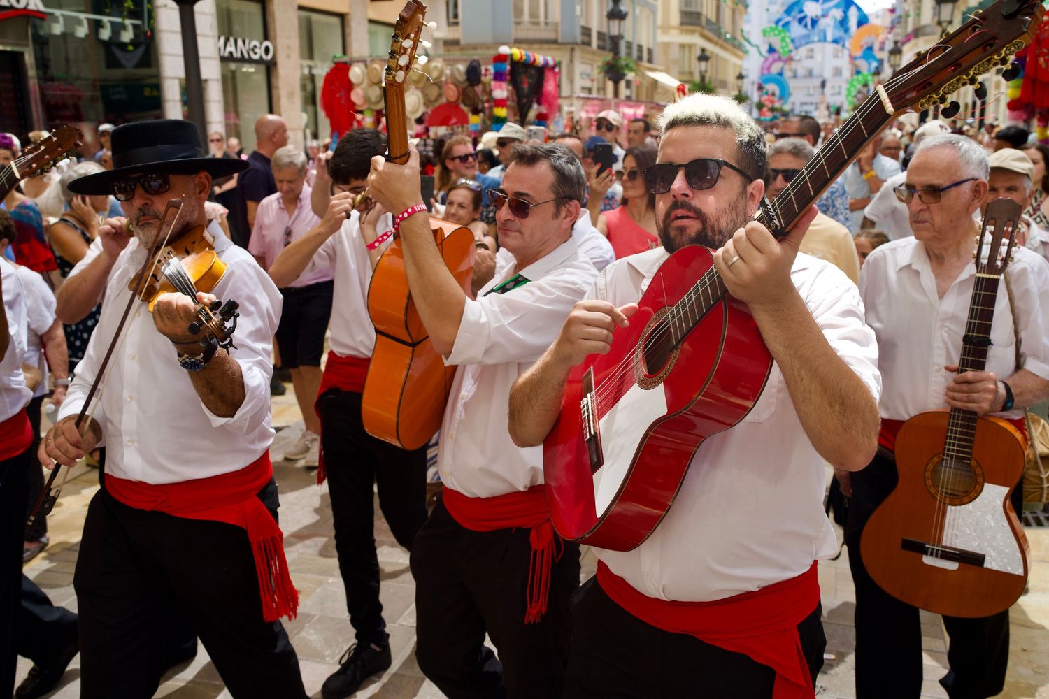 Verdiales musicians in traditional hats and red sashes playing guitars and violin in the crowded streets of Feria de Málaga.