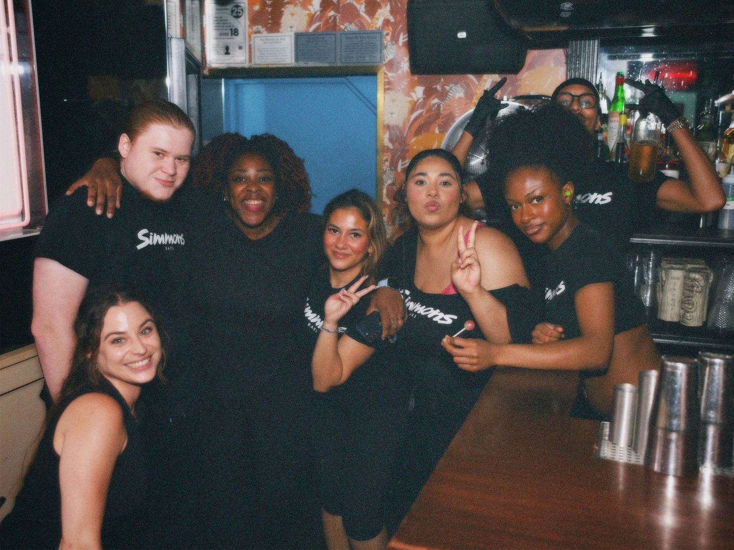 A group of Simmons bar staff posing and smiling together behind the bar counter. Commercial team portrait by ARIETA Studios.