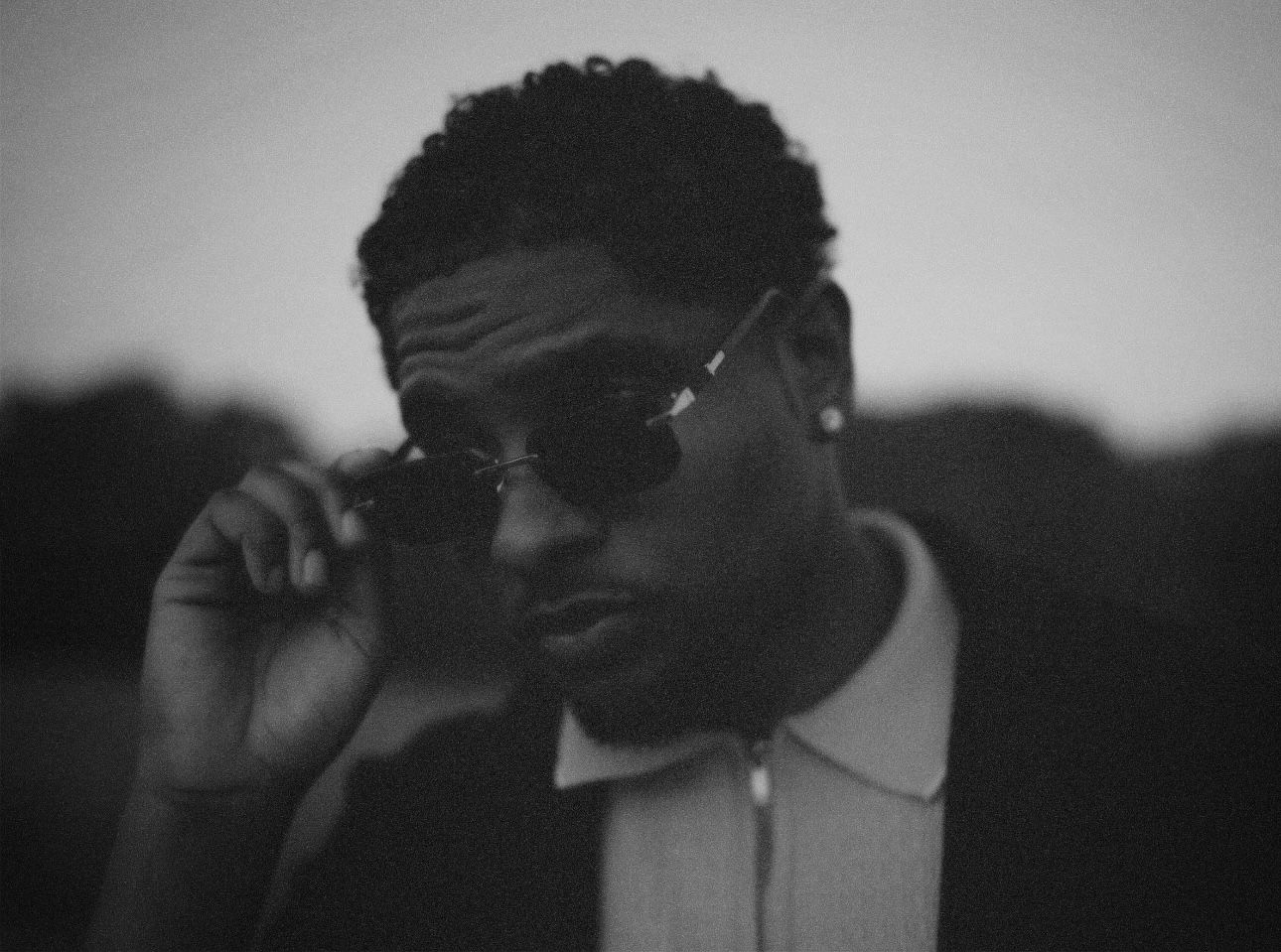 Cinematic black-and-white close-up of a man lowering his rimless sunglasses, looking directly into the camera lens with soft background blur