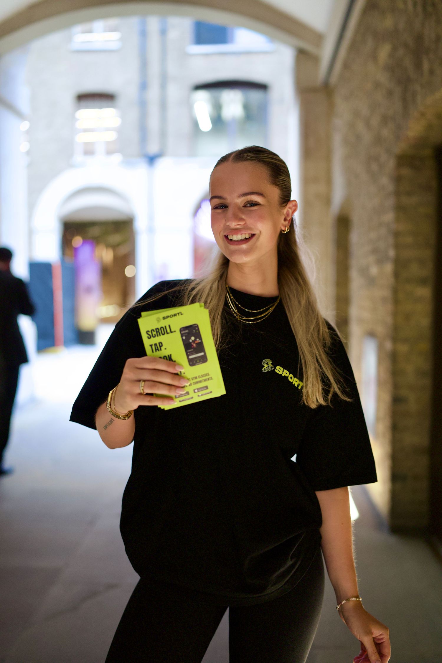 Smiling woman in a black SPORTL t-shirt holding a lime green event flyer at a London brand activation. Photo by ARIETA Studios