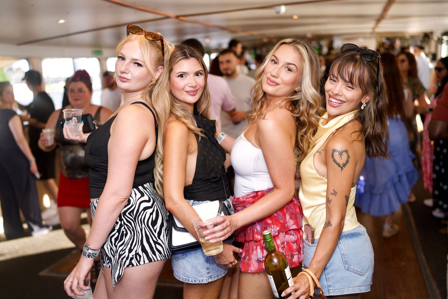 Four stylish women smiling and posing tightly together inside the deck of a busy London boat party.
