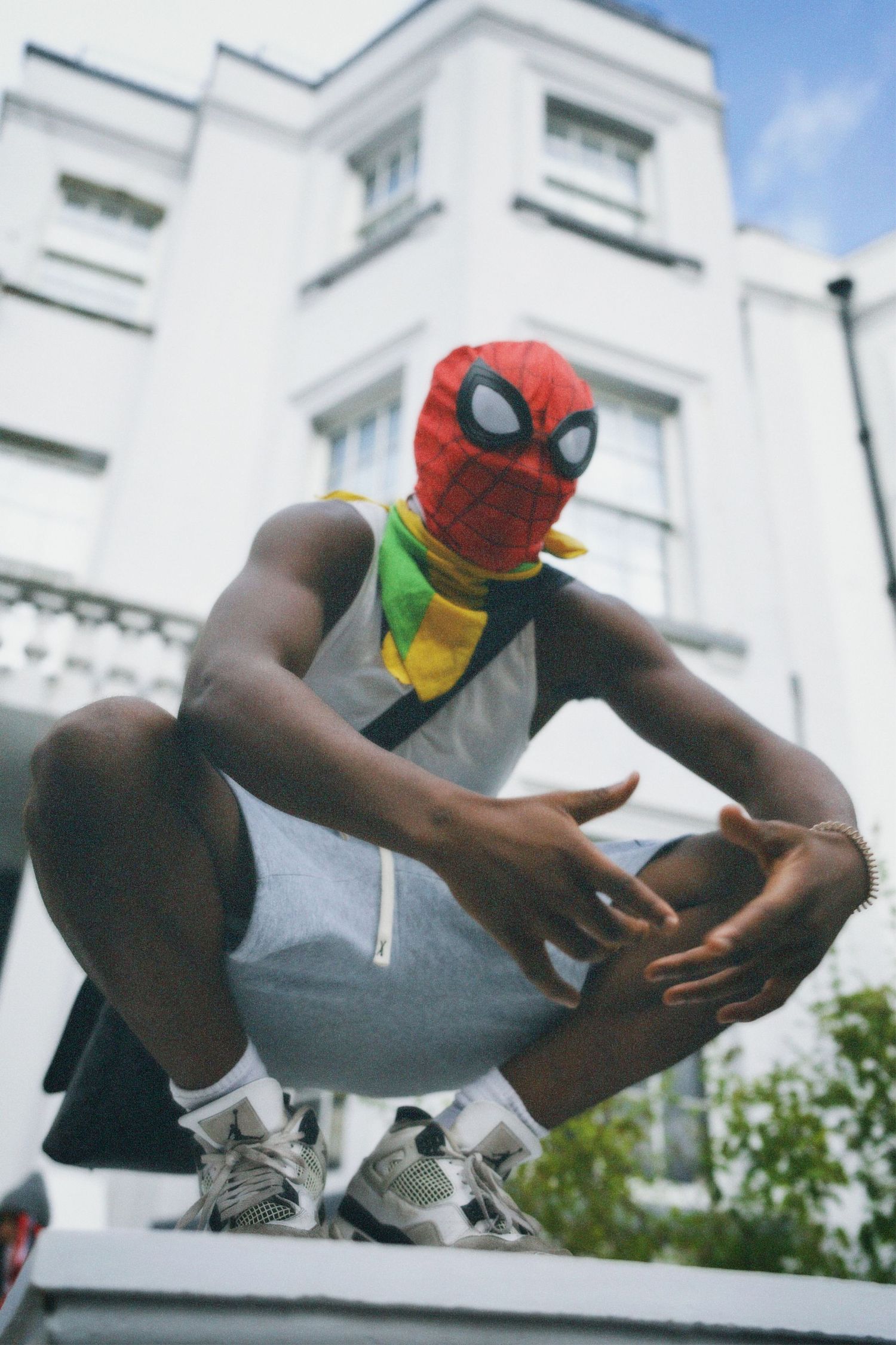 Editorial street fashion shot of a Spider-Man seated outdoors in front of leafy city trees.