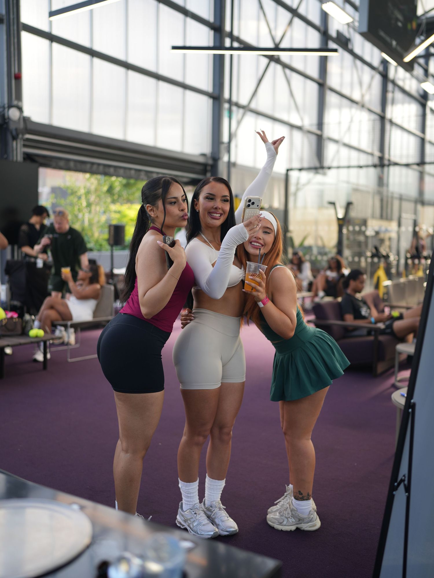 A candid courtside lifestyle photo of four women laughing and taking a selfie together in coordinated summer sports clothing.