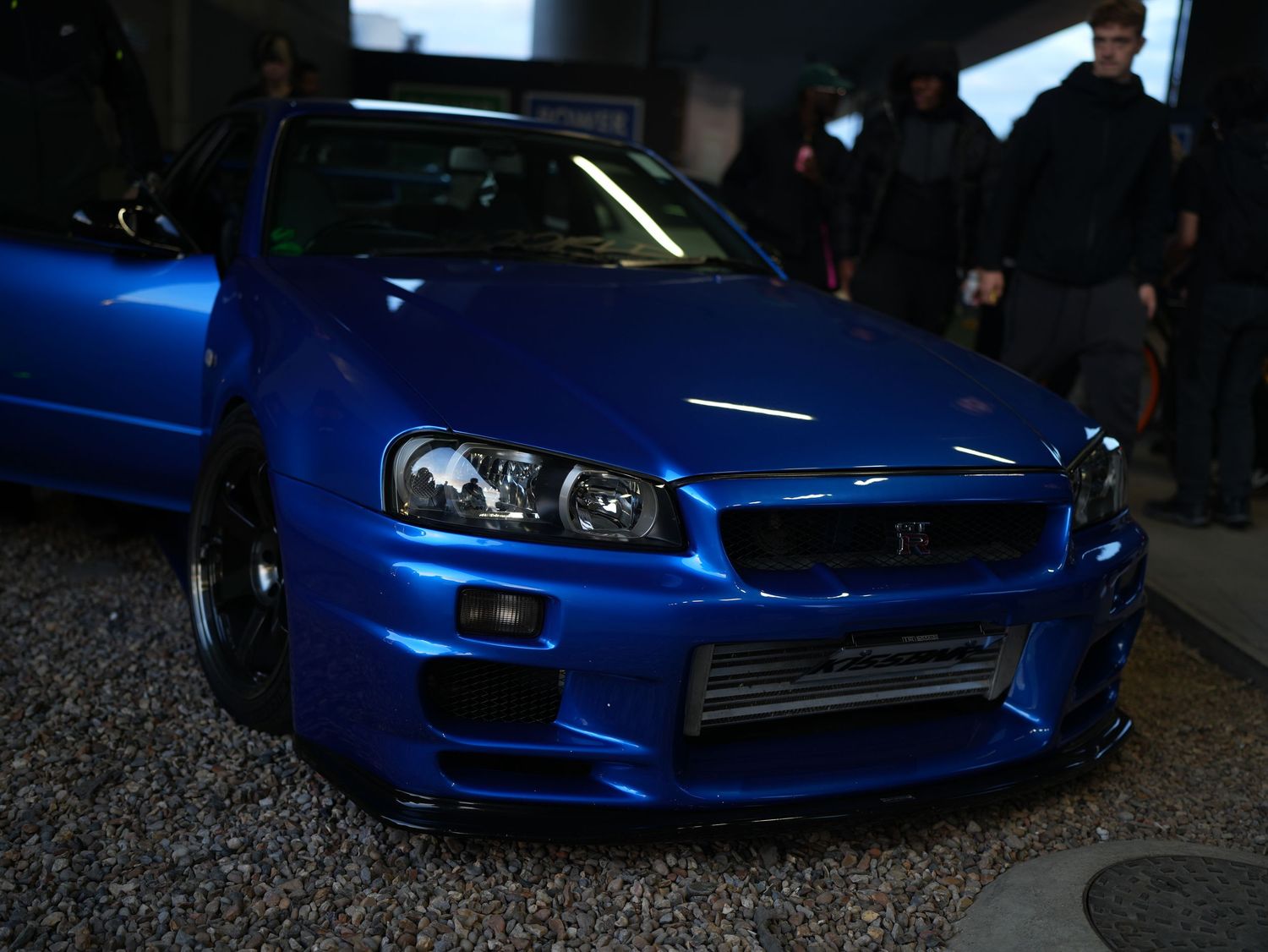 A modified bayside blue Nissan Skyline GT-R R34 parked at an exclusive late-night automotive event in London.