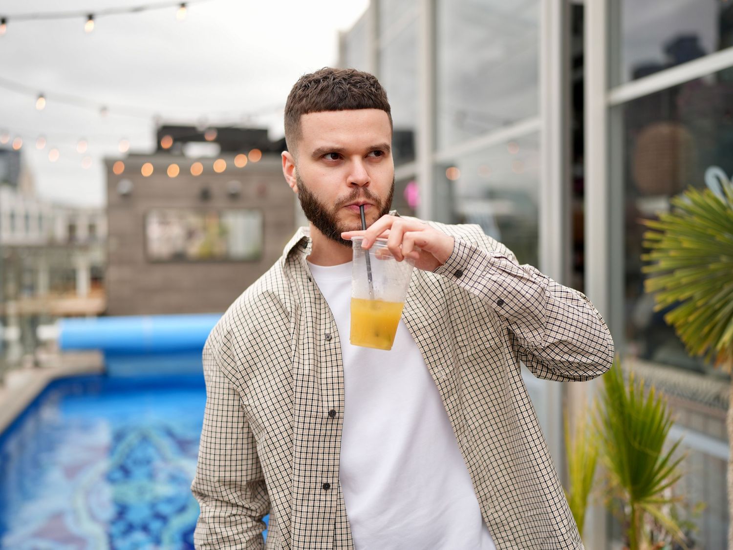 A man in a patterned shirt sipping a drink by an open-air poolside deck.