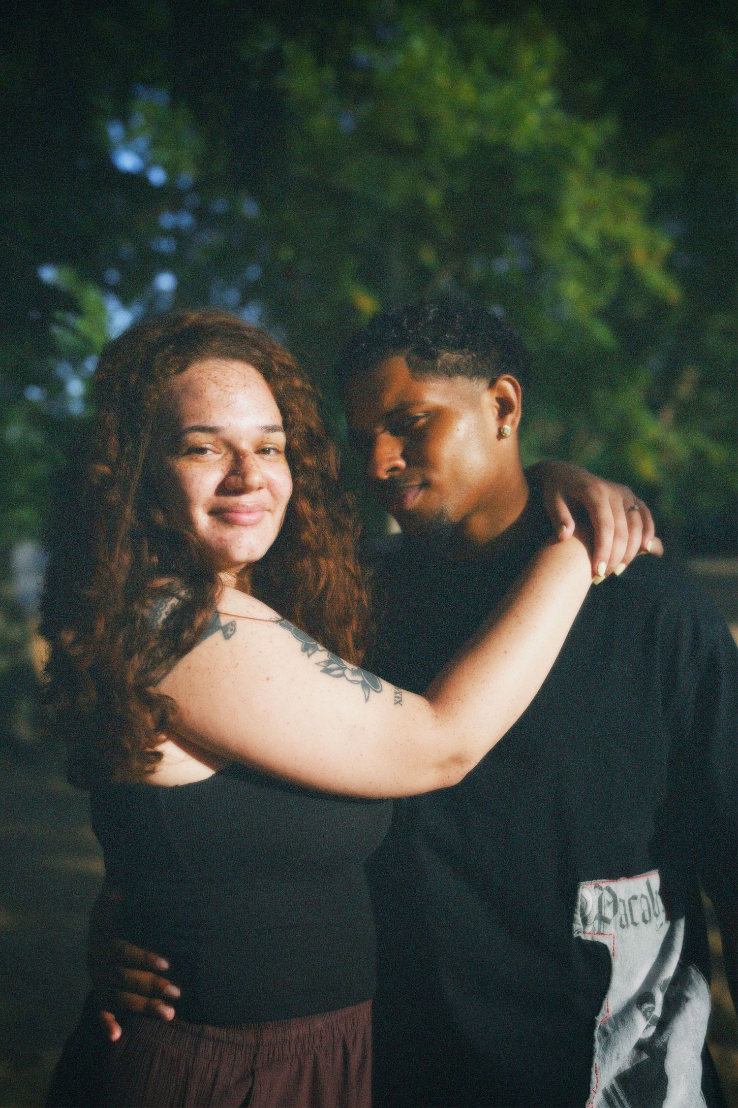 Intimate close-up portrait of a couple outdoors, with the woman wrapping her arm around the man's shoulders under warm golden sunlight.