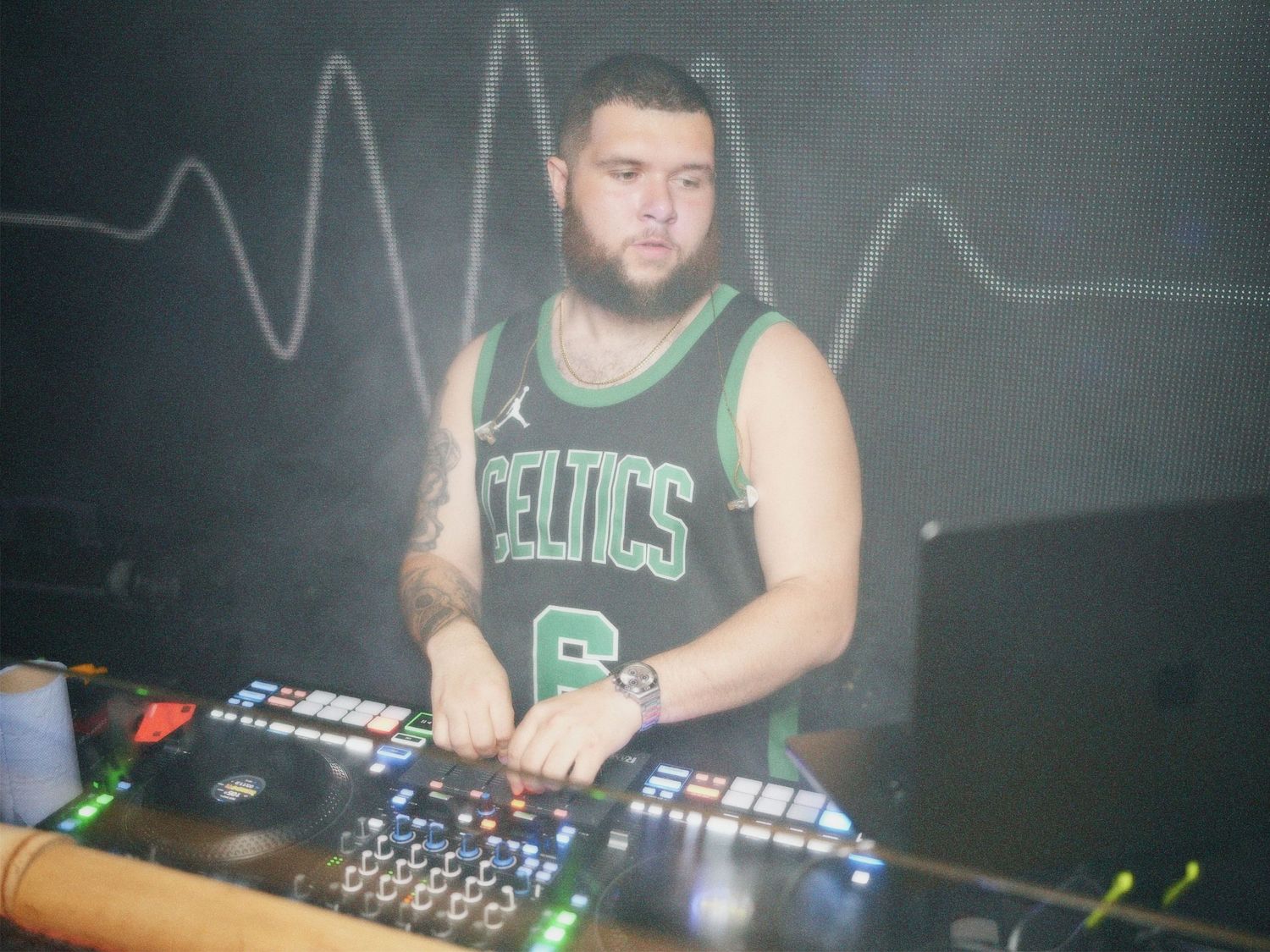 DJ wearing green vest mixing set behind decks at venue event by ARIETA Studios.