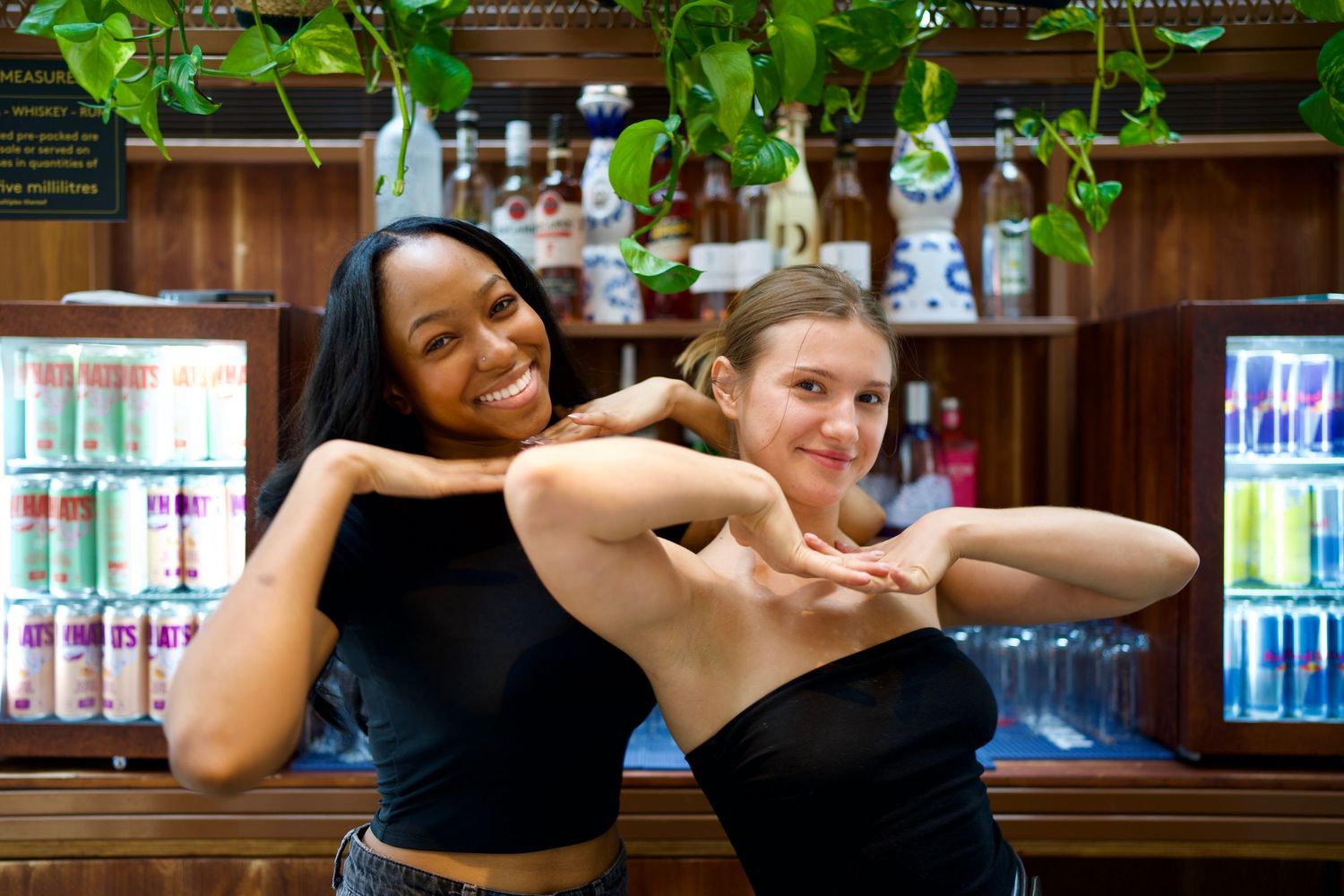 Two smiling women posing playfully together in front of a bar at a high-energy nightlife event. Photo by ARIETA Studios.