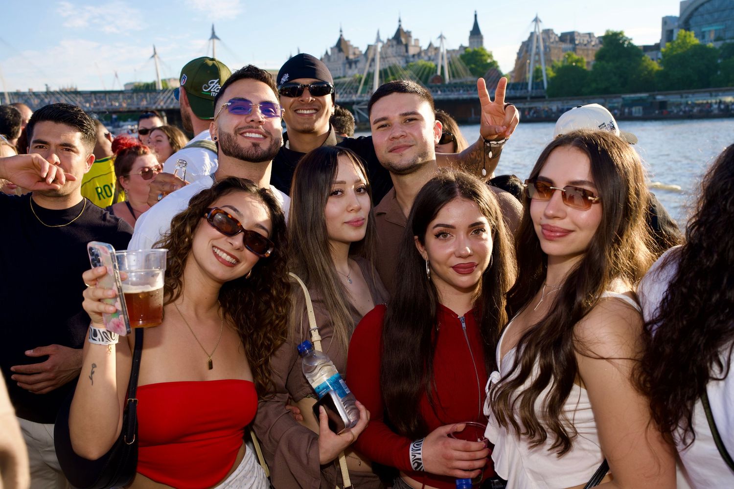 A large, diverse group of friends holding drinks and throwing up peace signs outside on the boat deck.