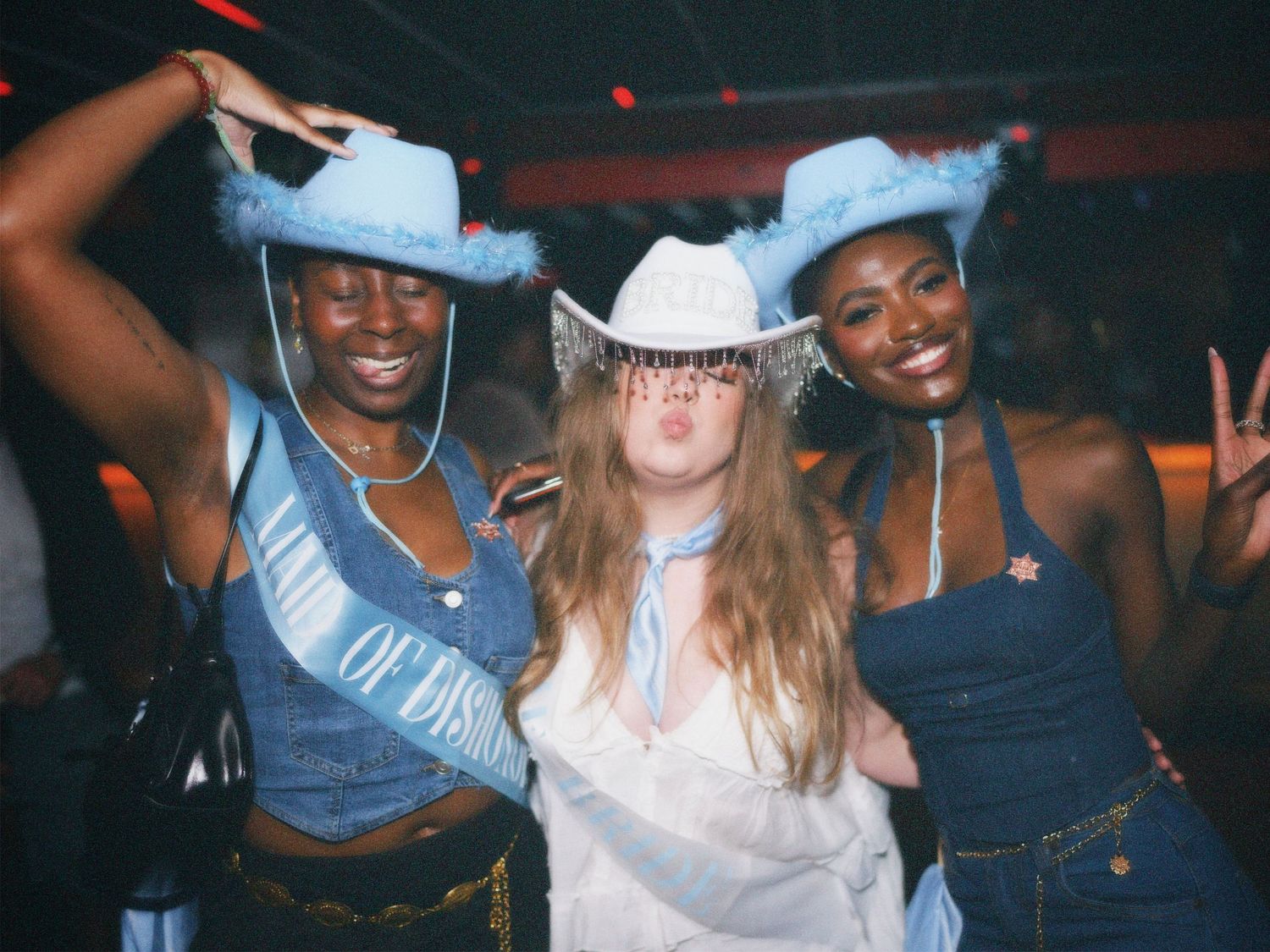 Three women wearing blue cowboy hats smiling and posing together at a themed nightlife event. Photo by ARIETA Studios.