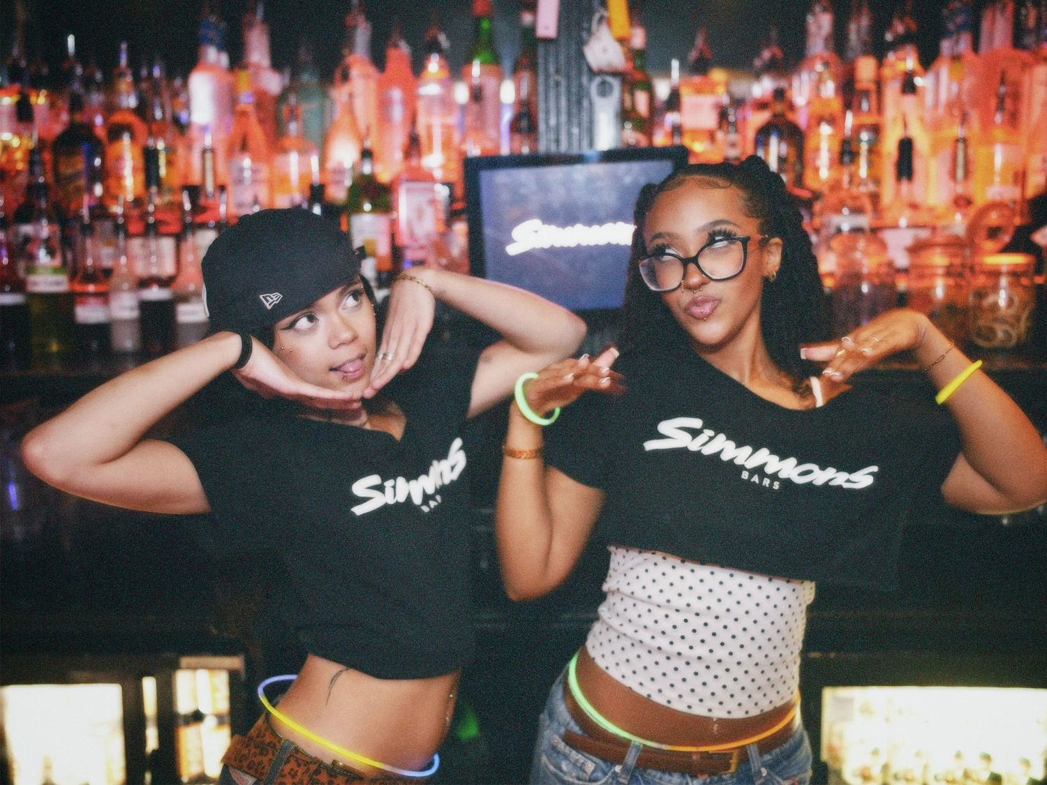 Two women in black branded crop tops posing in front of lit bar shelves by ARIETA Studios
