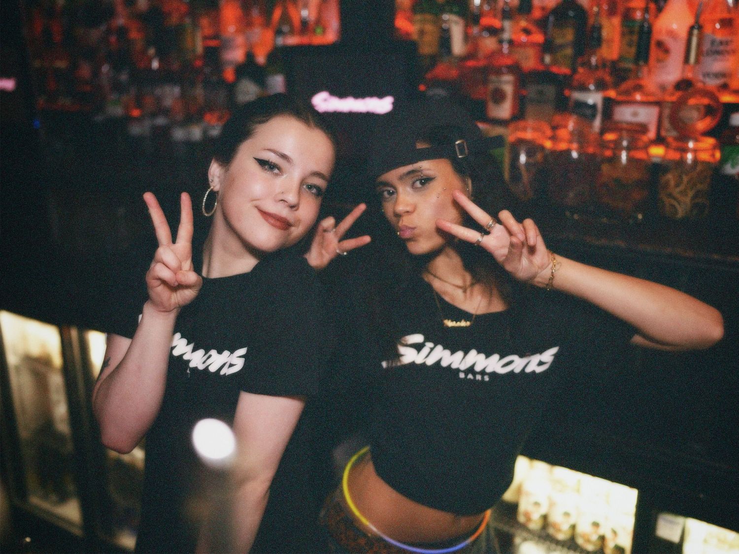 Two women in matching black tops throwing peace signs behind bar counter by ARIETA Studios.