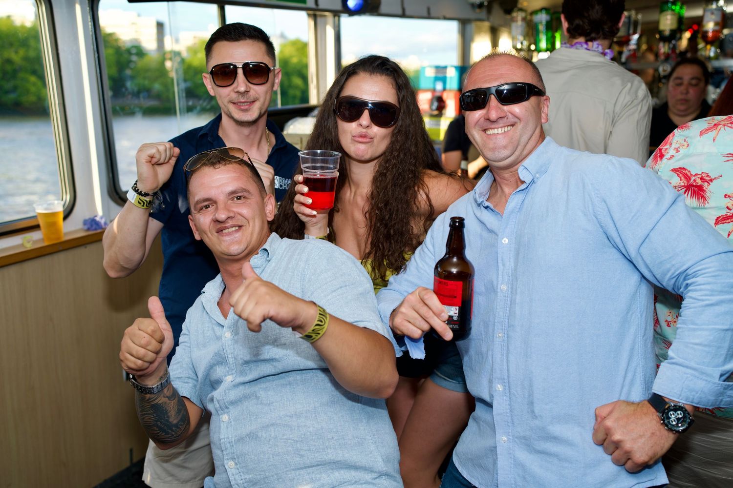 Group of friends raising beer bottles and cheering on River Thames boat party by ARIETA Studios.