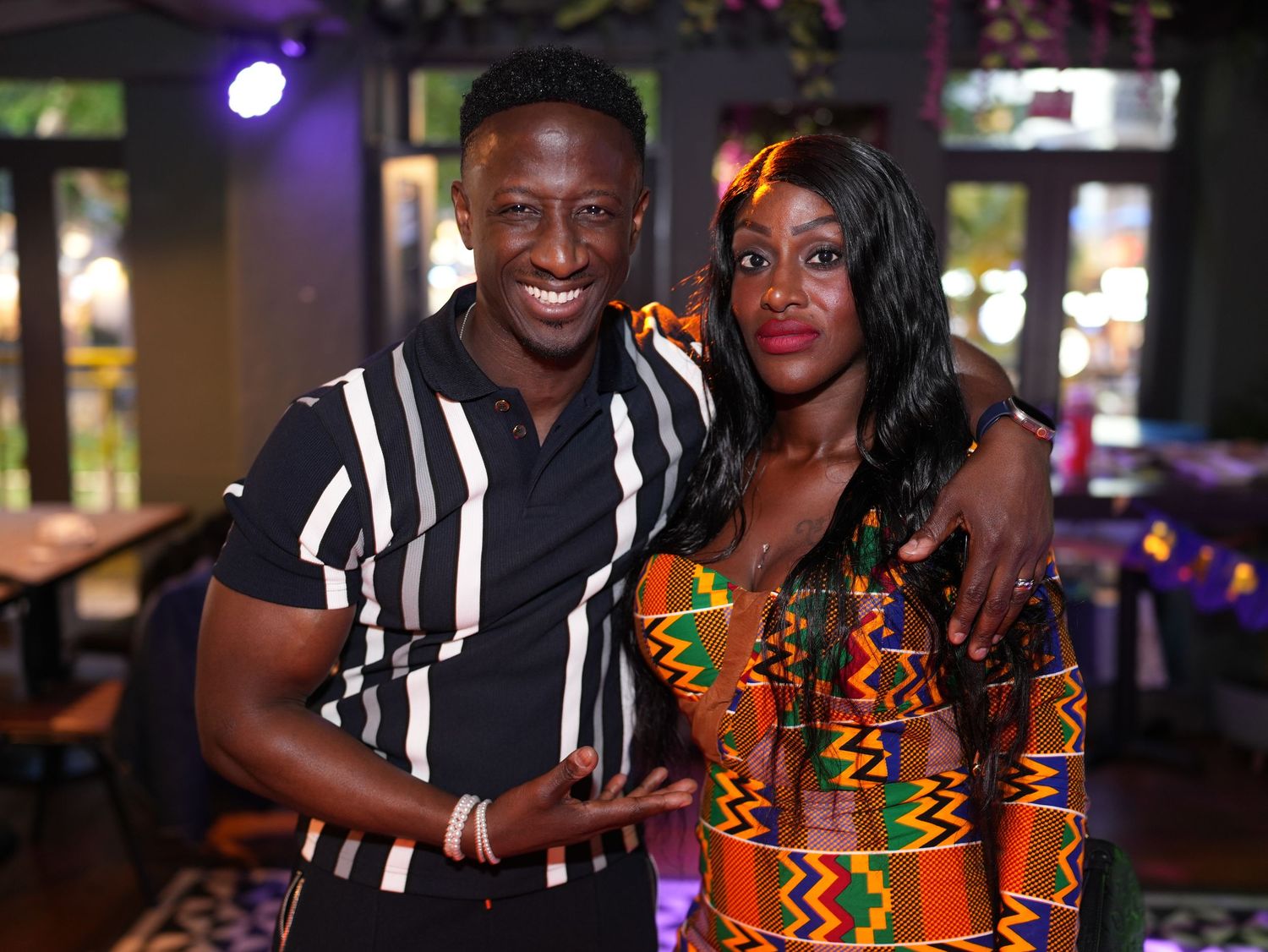 A man in a striped polo and a woman in a patterned dress posing together at an evening event.