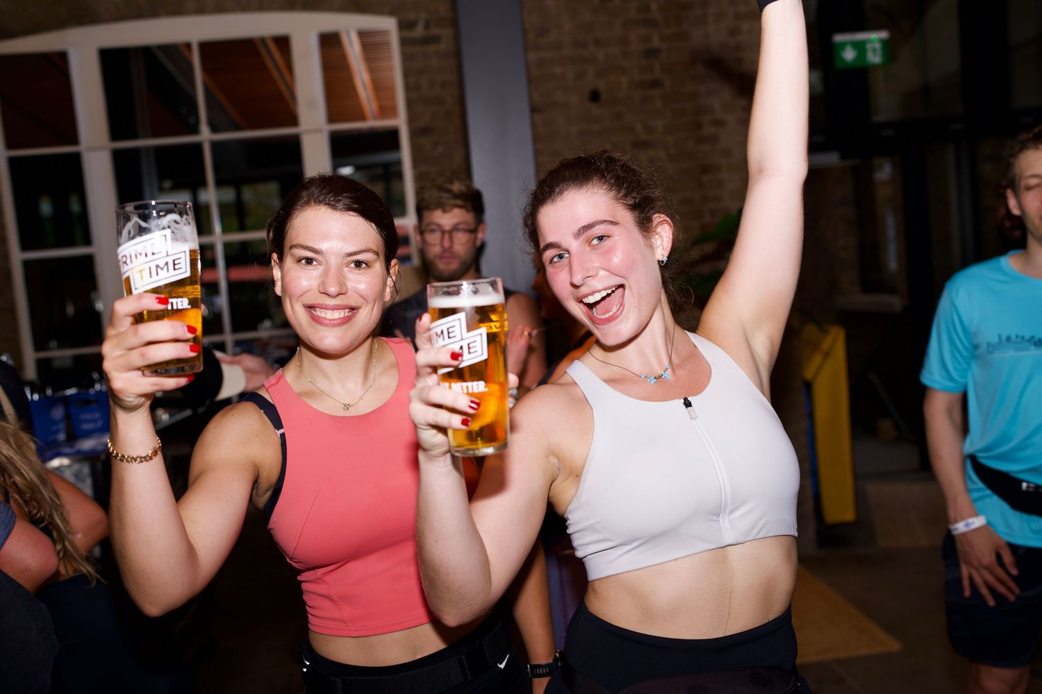 Two smiling women celebrating and raising pints of beer at an energetic indoor event after-party. Captured by ARIETA Studios.