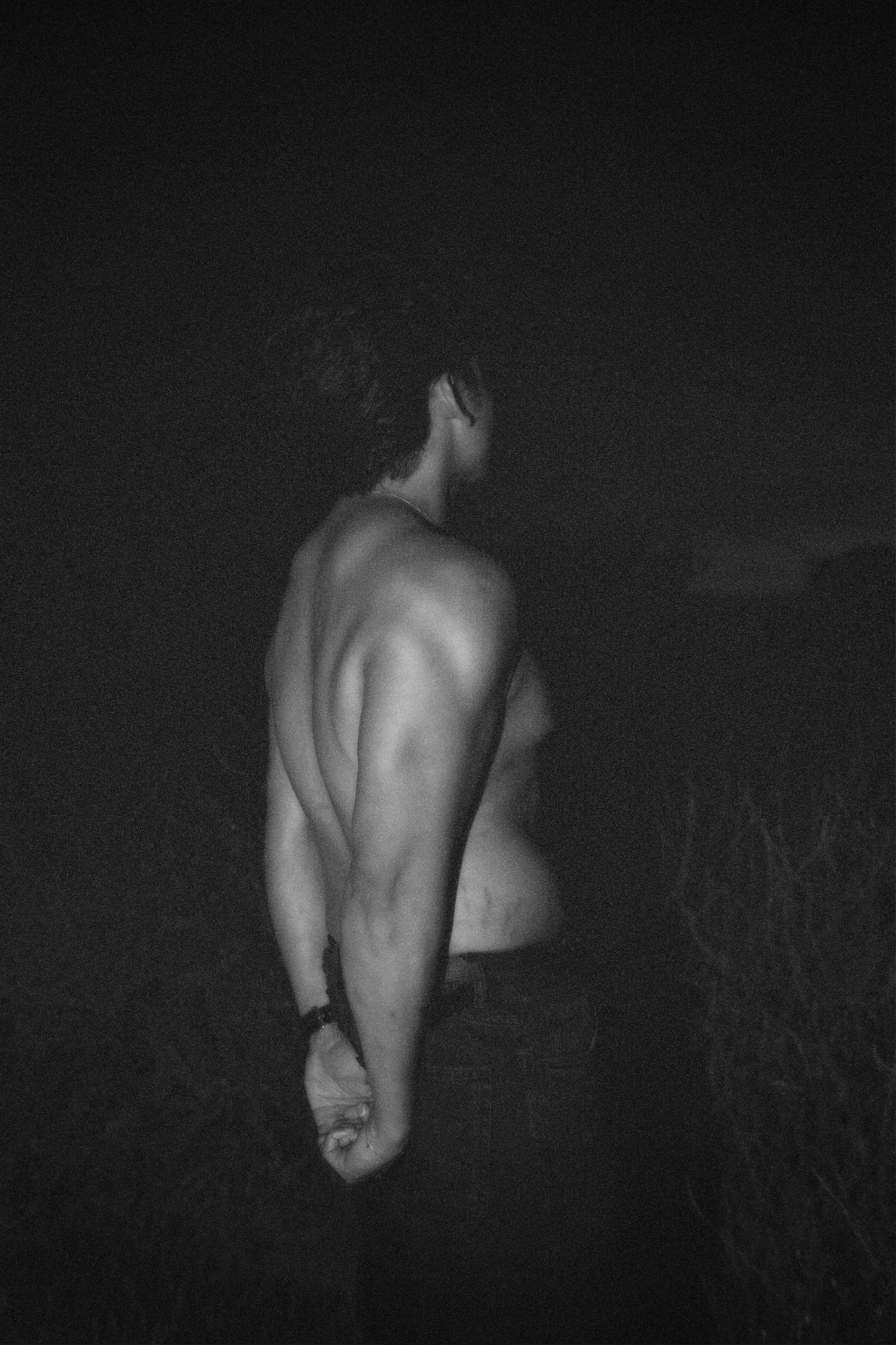 Gritty black-and-white shot of a Alastair shirtless from behind with arms clasped, showcasing back definition against a dark background.