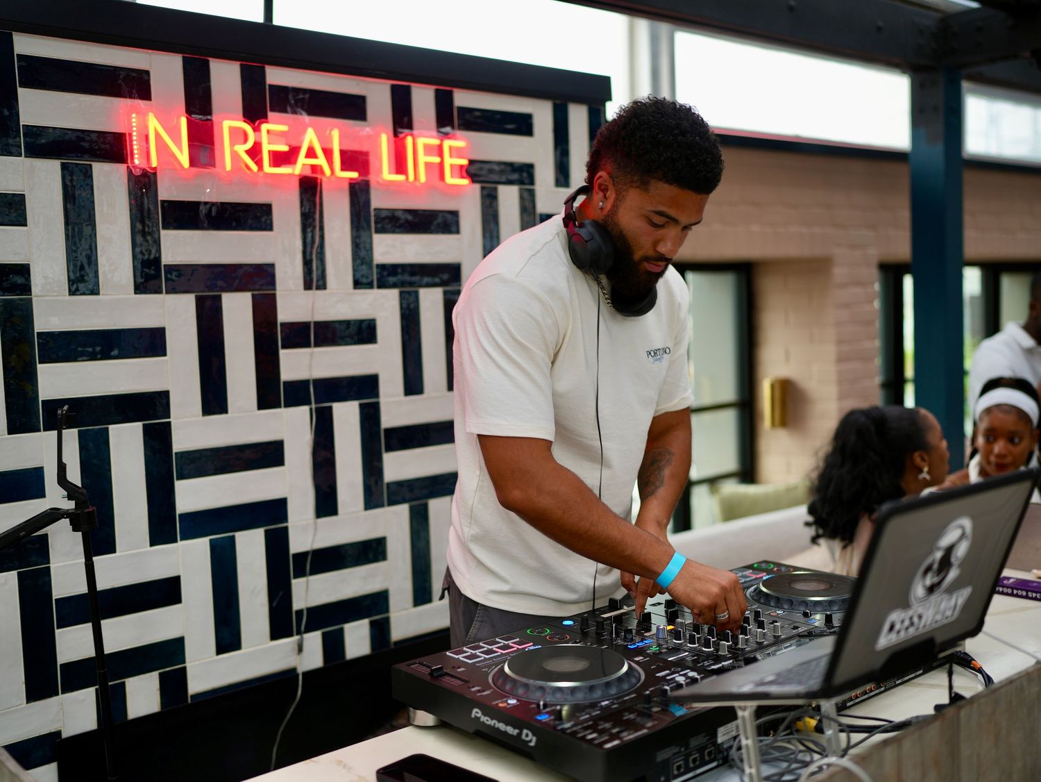 A DJ mixing tracks at a premium rooftop event next to a red neon sign reading 