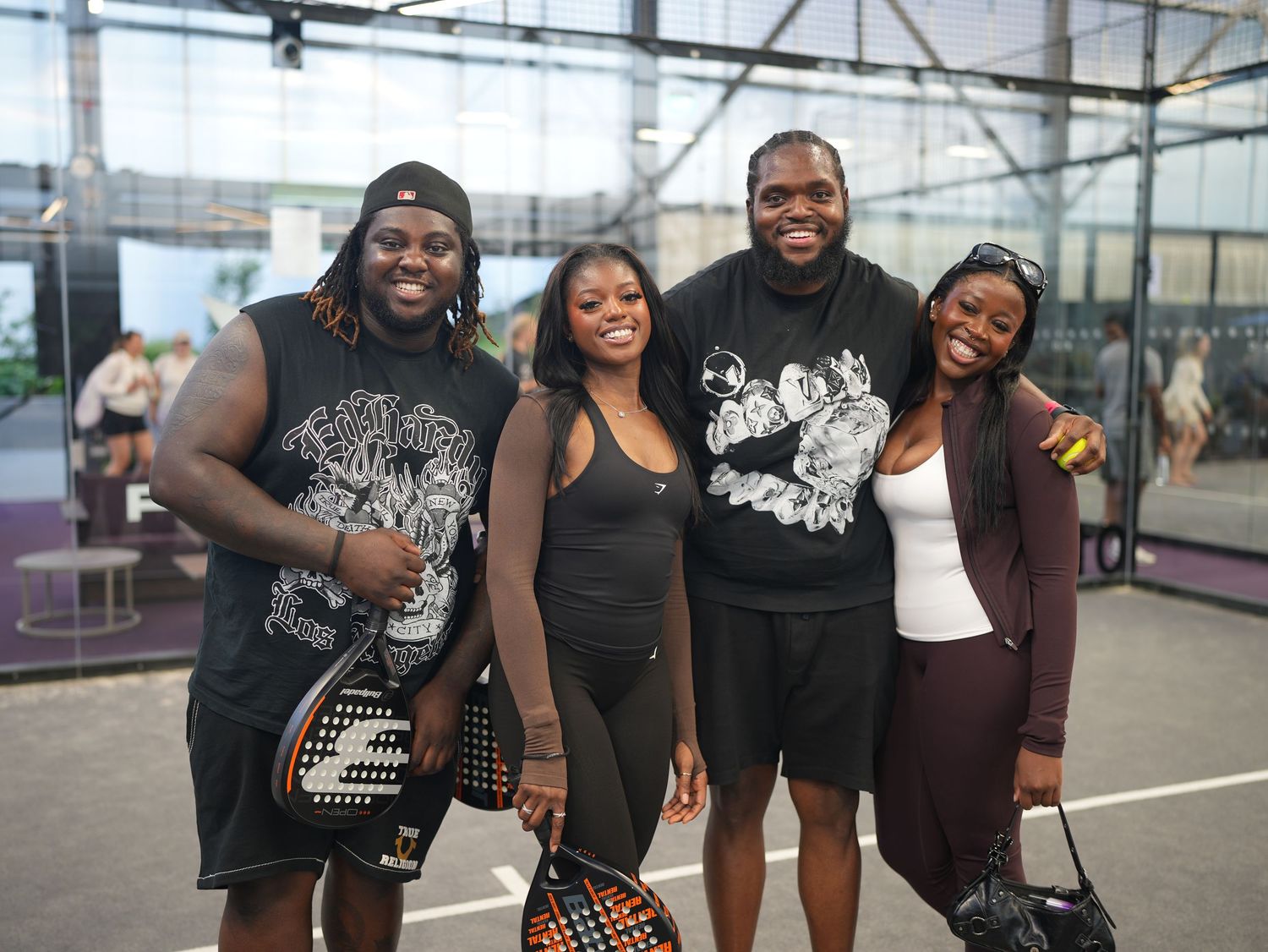 A group portrait of two men and two women smiling together on an indoor padel court, dressed in stylish black and dark activewear.