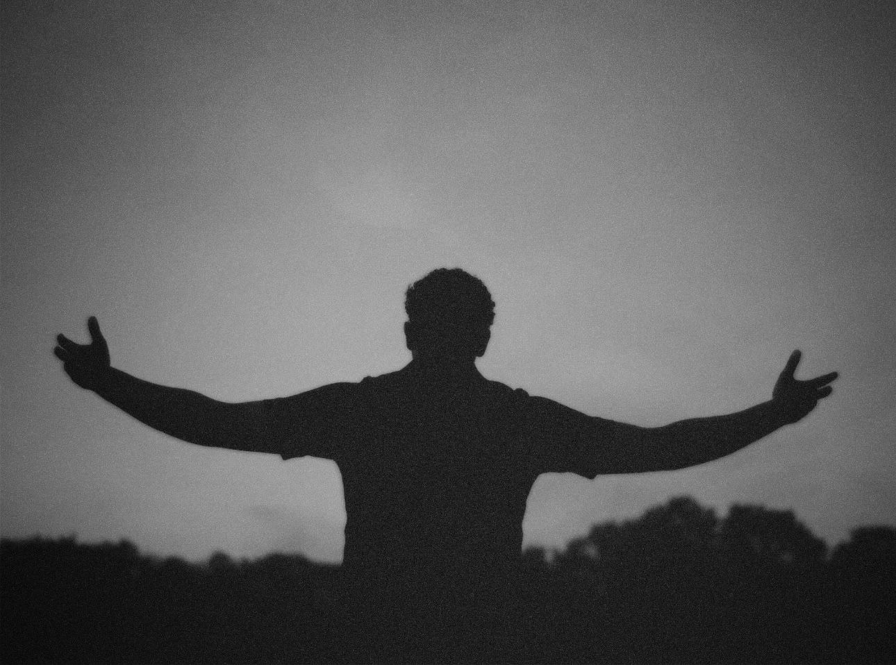 Grainy black-and-white silhouette of a man standing outdoors at dusk with his arms open wide against the low-light sky.