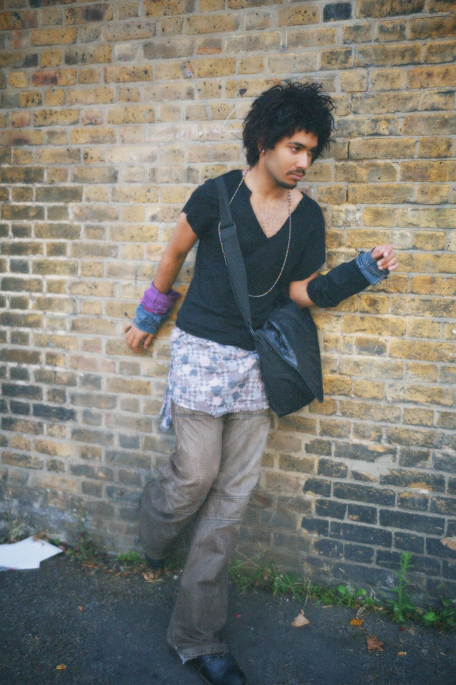 Stylish young man with a crossbody bag posing casually against an East London brick wall in a vintage outfit