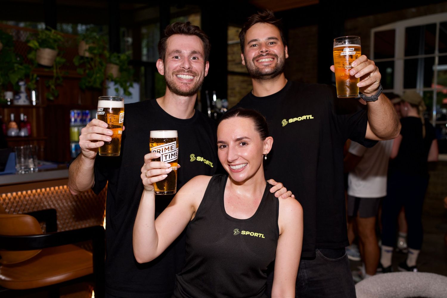 Two men and a woman in SPORTL gear smiling and raising pints of Prime Time Beer at a London venue event. Photo by ARIETA Studios.