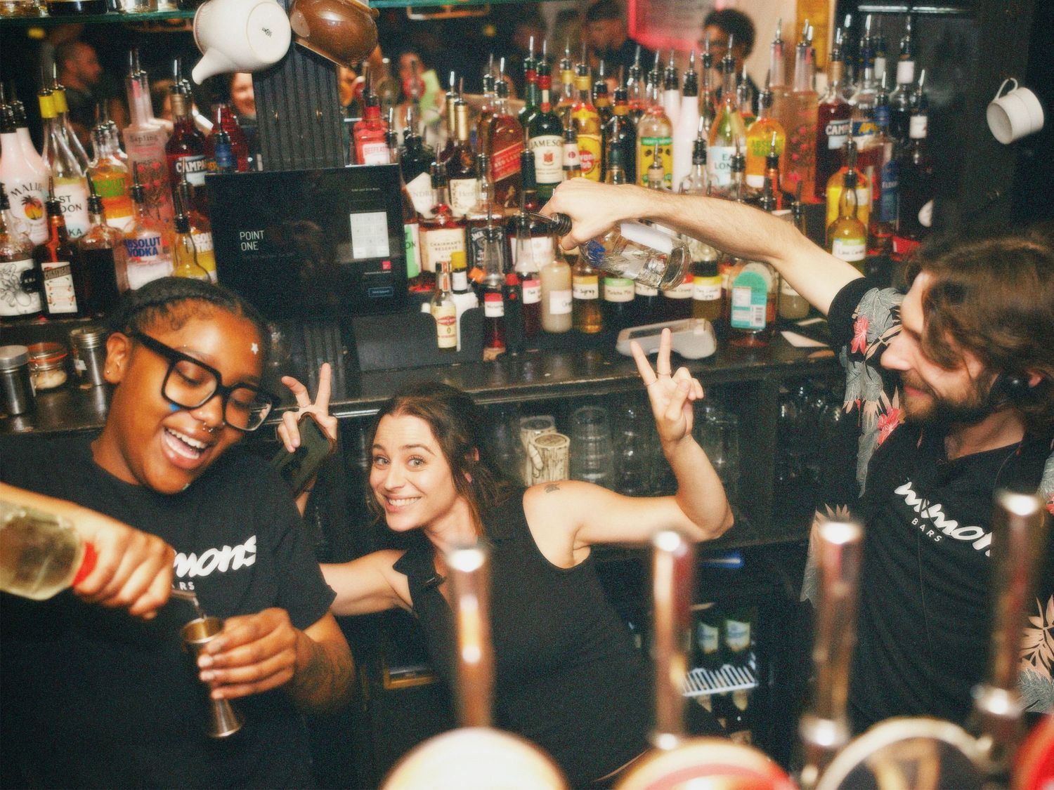 Bartenders smiling and pouring drinks behind a fully stocked bar at Simmons Shoreditch. Nightlife photography by ARIETA Studios.