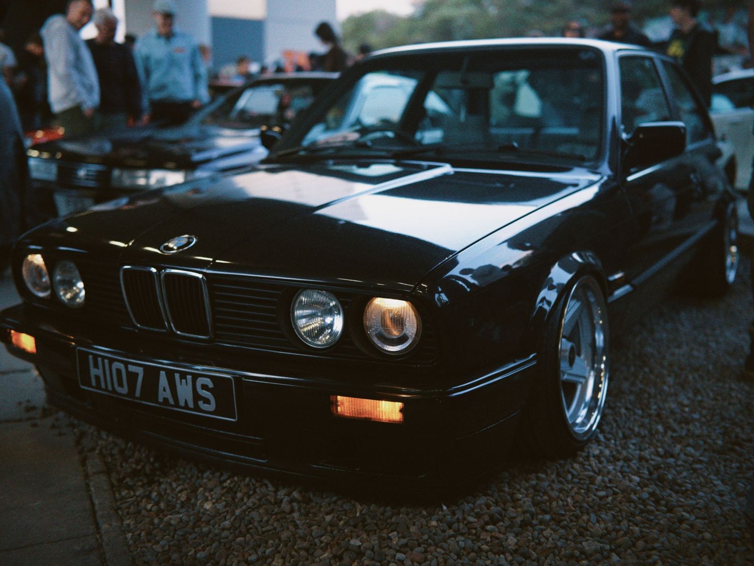 Front three-quarter view of a modified black classic BMW E30 saloon on deep dish wheels.