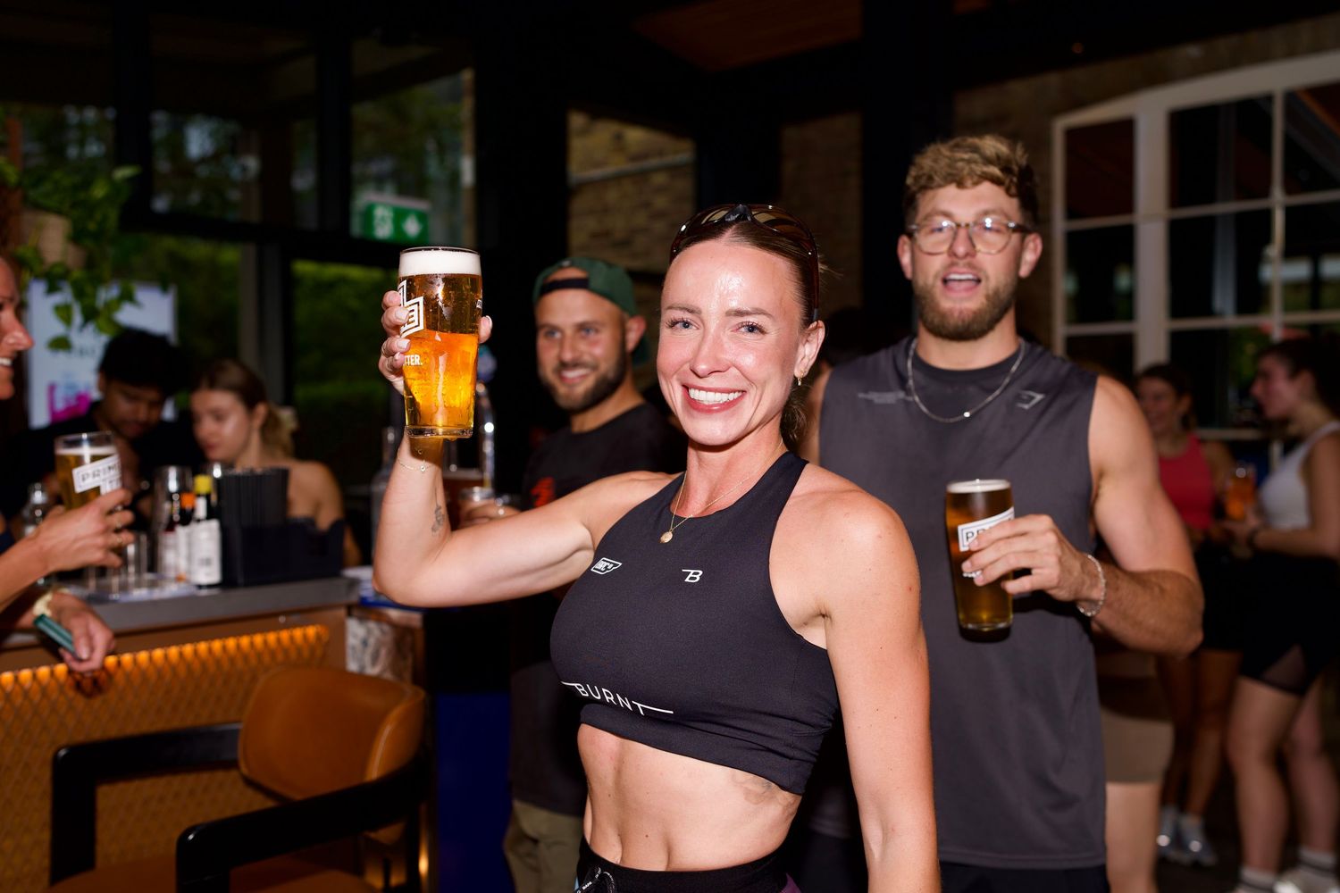 Smiling woman in a black sports top raising a pint of beer at a lively indoor bar activation. Commercial photography by ARIETA Studios.