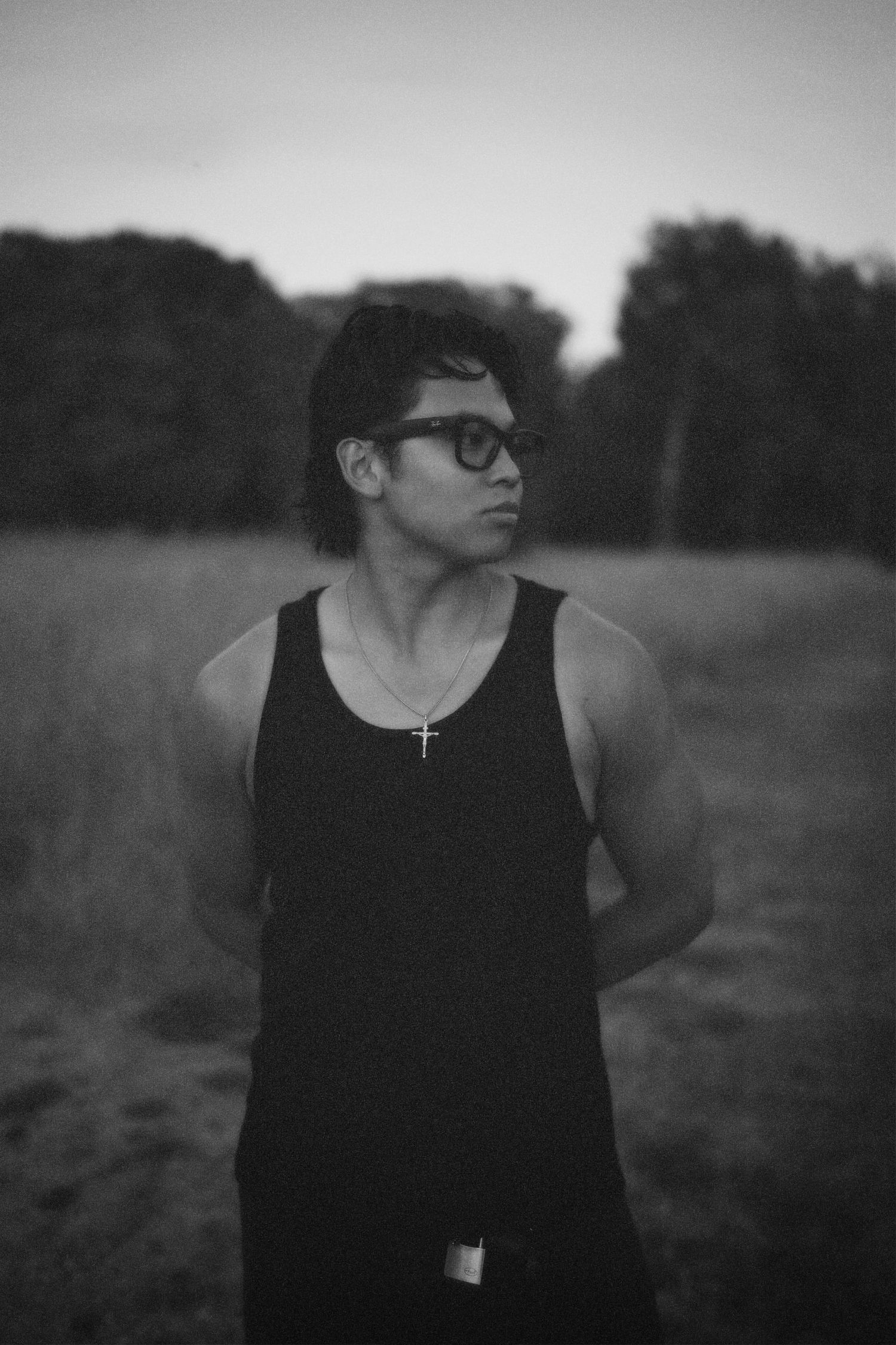 Grainy black-and-white portrait of Alastair with glasses looking sideways at dusk, wearing a tank top against a dark, field-like background.
