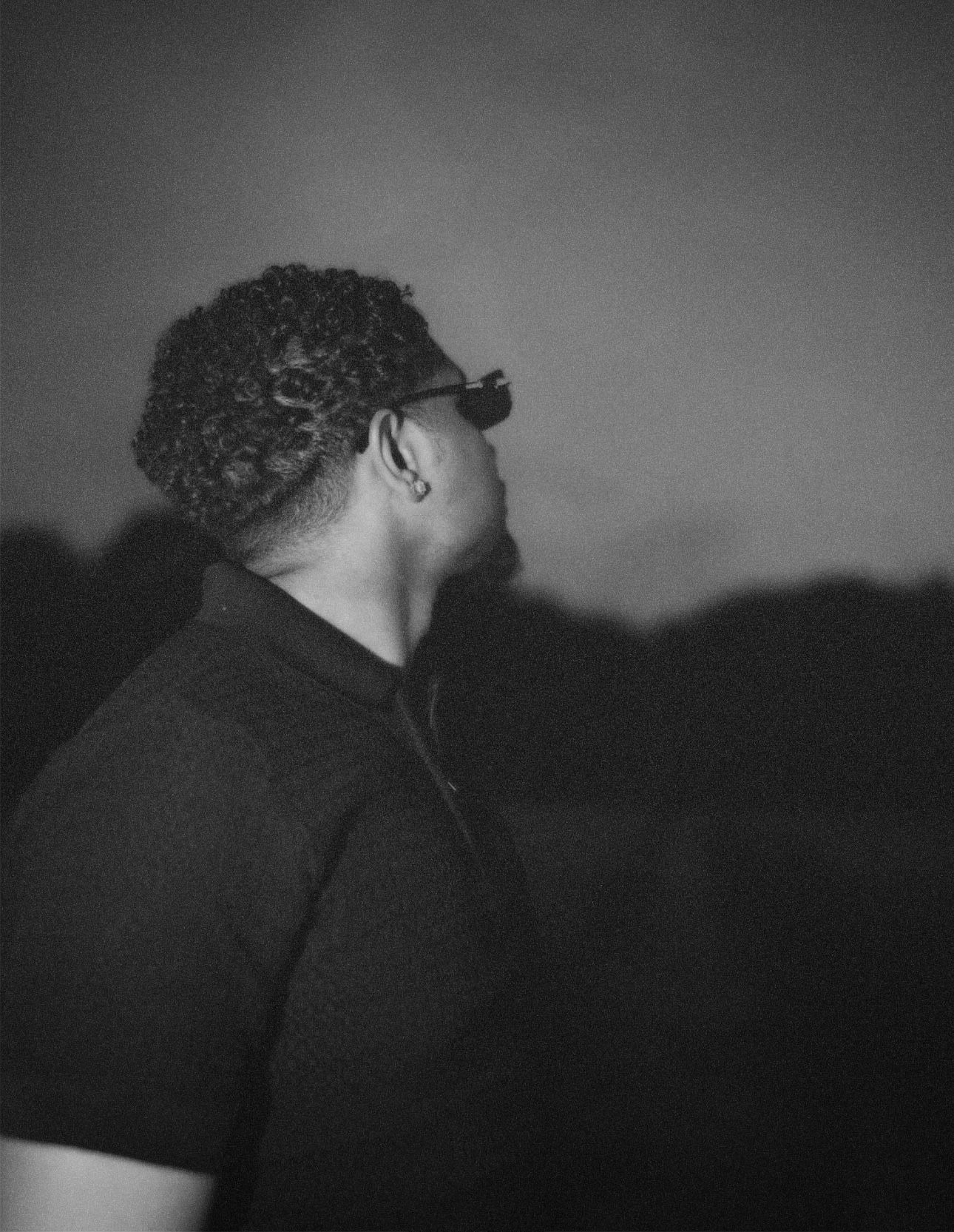 Grainy black-and-white profile shot of a man in a black polo shirt and sunglasses looking upward against a dark evening horizon.
