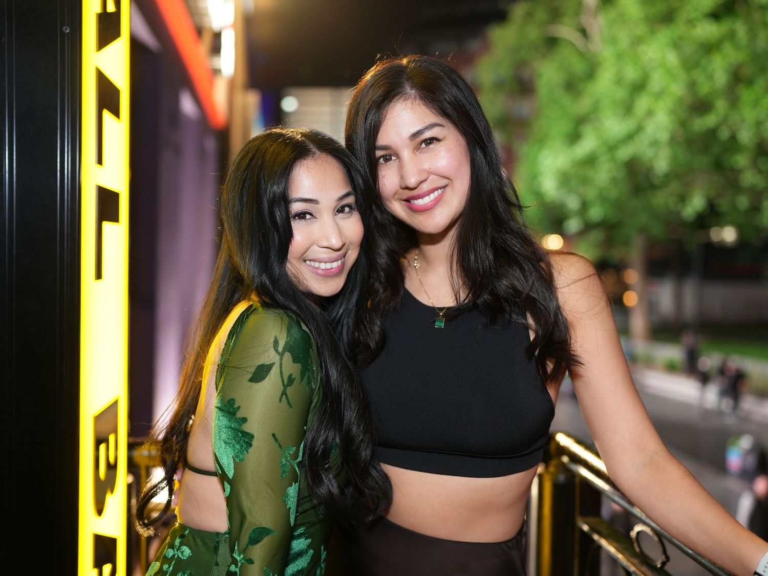 Rehana in her green dress smiling and hugging a friend in a black crop top near a window.