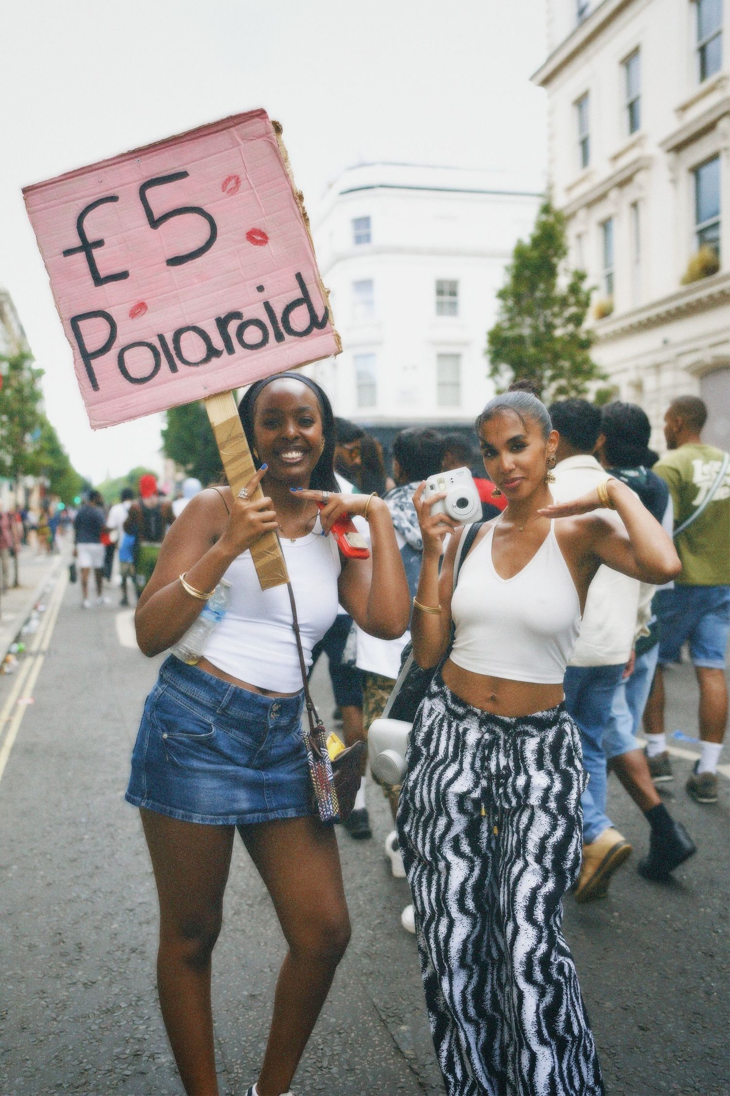 Two attendees posing on the road next to a vendor holding a £5 Polaroid sign.