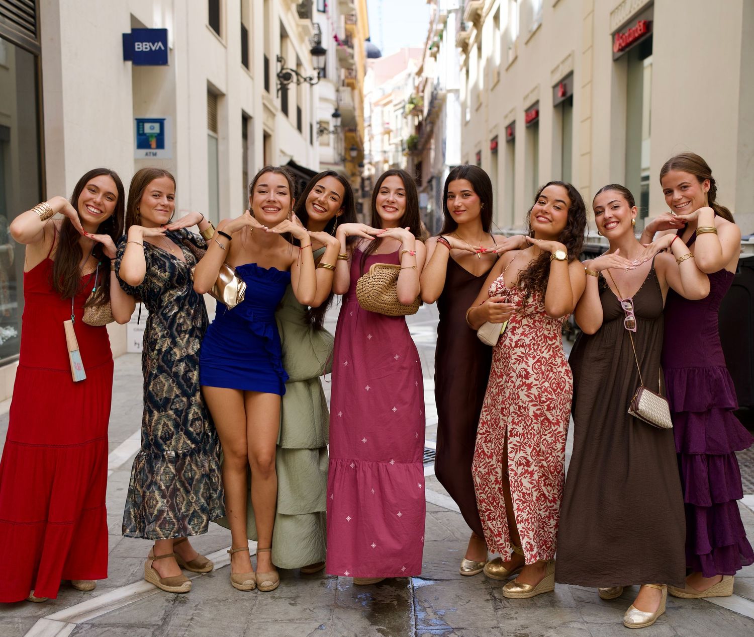Group of nine friends in colorful summer dresses posing with cheerful smiles in a sunlit street at Feria de Málaga.