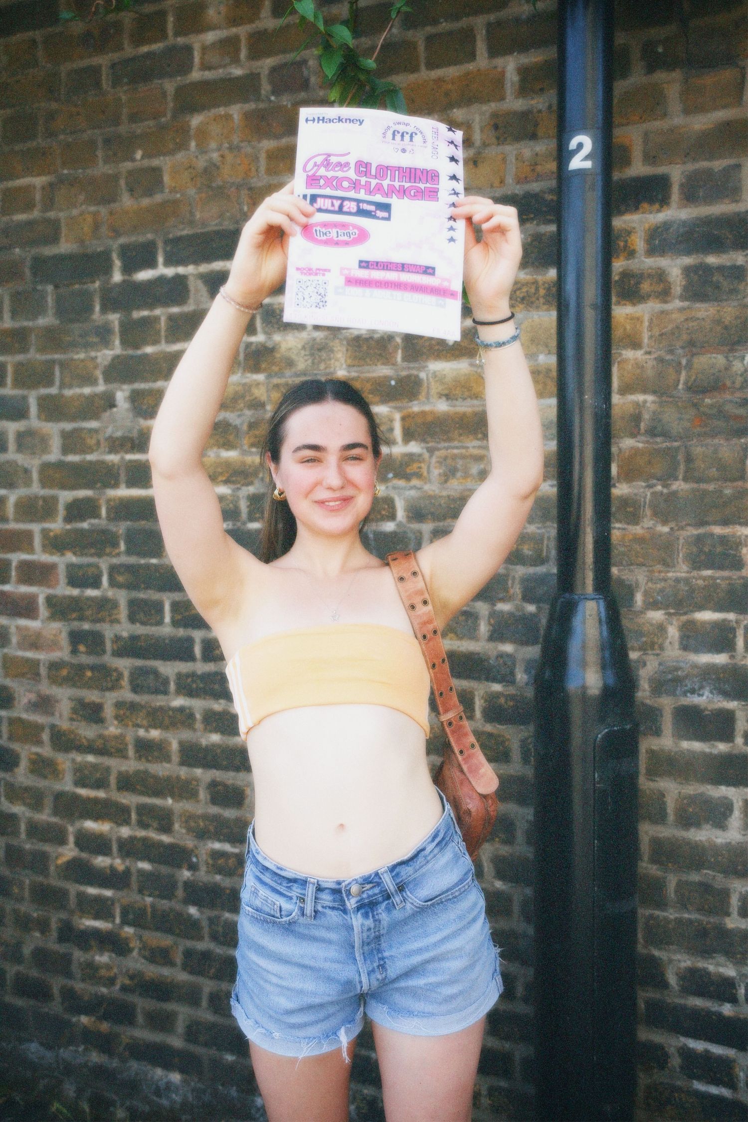 Smiling woman holding up a Fashion For Future event flyer against a brick wall outside in Hackney