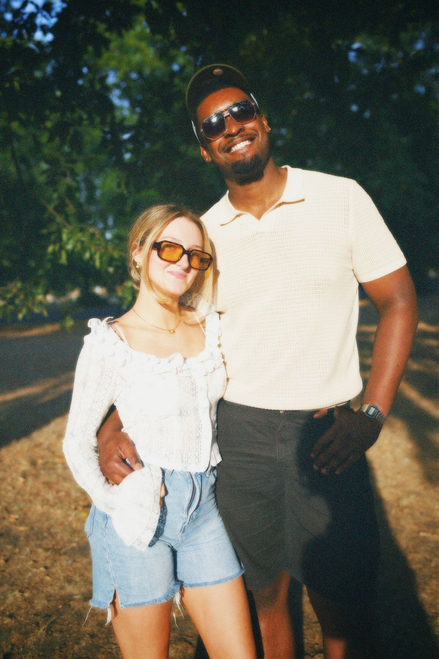 A stylish couple wearing sunglasses standing together in a park setting, smiling into the camera with soft summer light in the background.