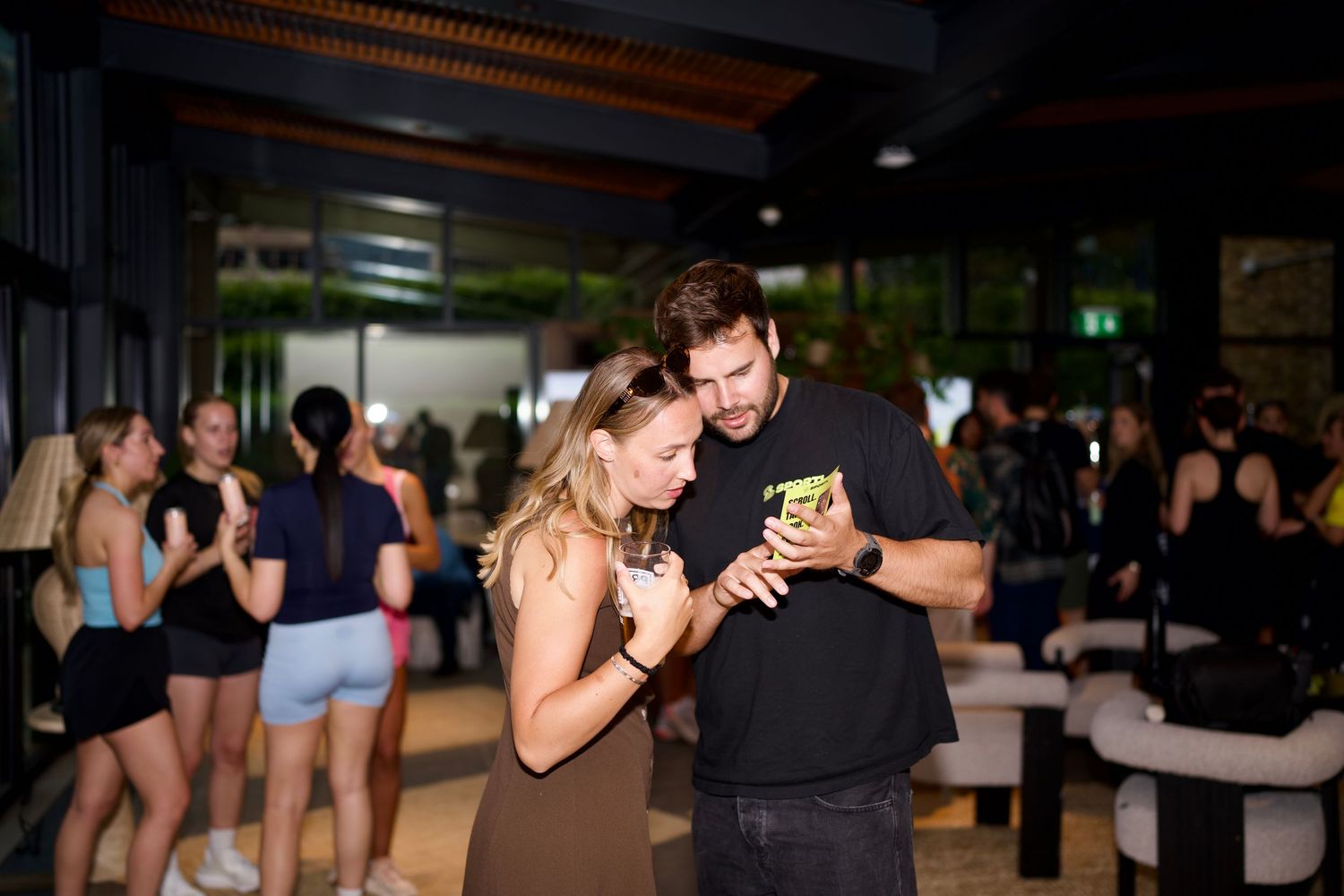 A man in a SPORTL t-shirt showing a phone screen to a woman during a lively commercial event after-party. Photo by ARIETA Studios.